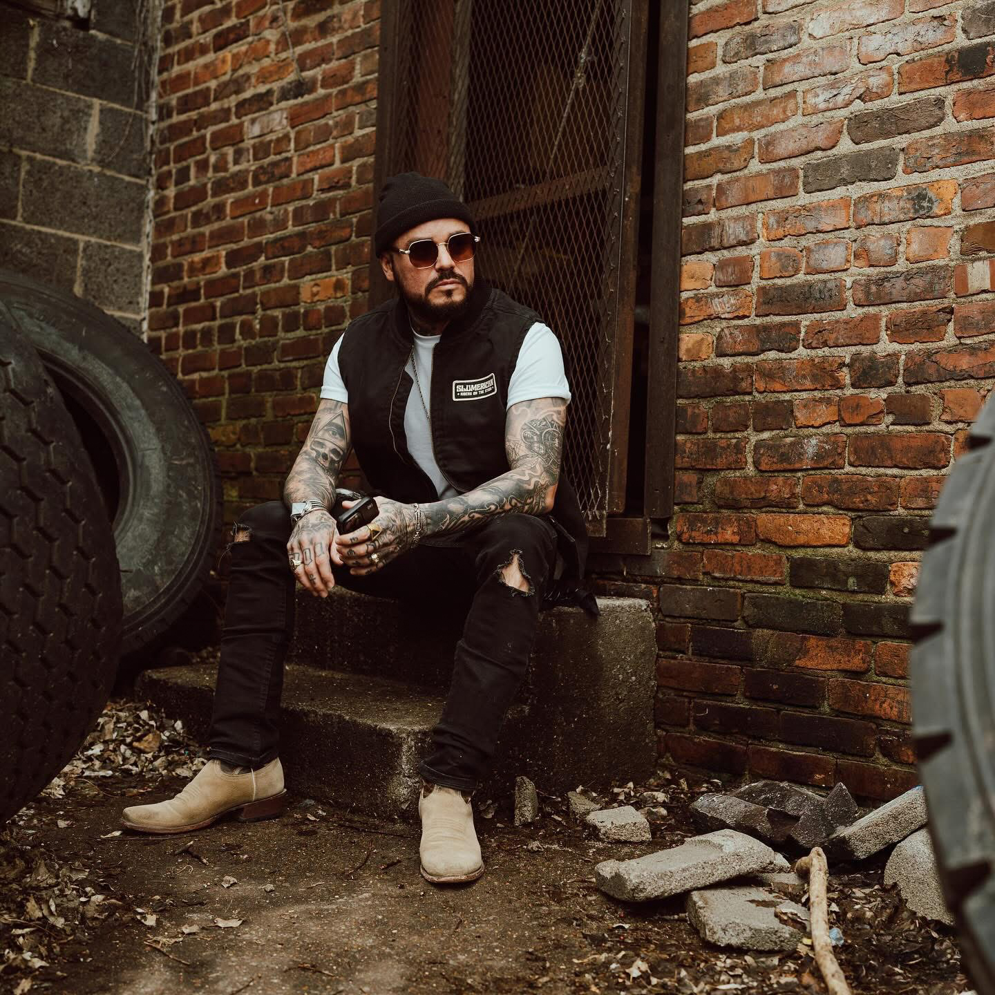Man with tattoos, beard, sunglasses, black beanie, black vest, white shirt, black ripped jeans, and beige shoes sitting on steps outside brick building holding a drink.