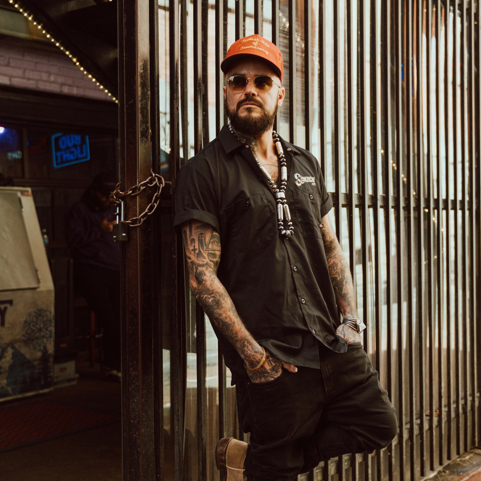 Man with tattoos, beard, sunglasses, orange cap, black shirt, and black pants standing outside near metal bars.