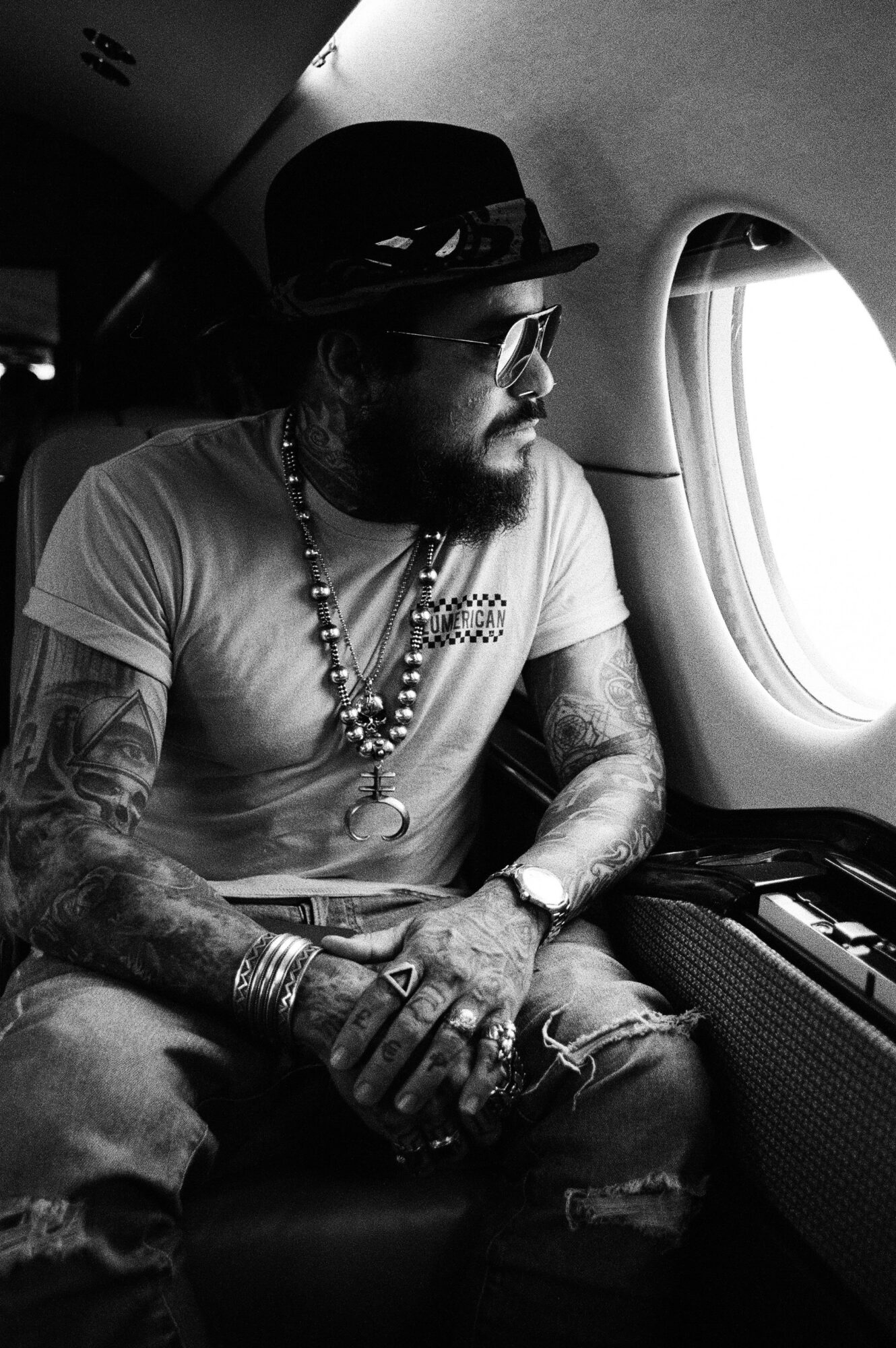 Man with tattoos, wearing a hat and sunglasses, sitting by airplane window, holding a phone, in black and white.