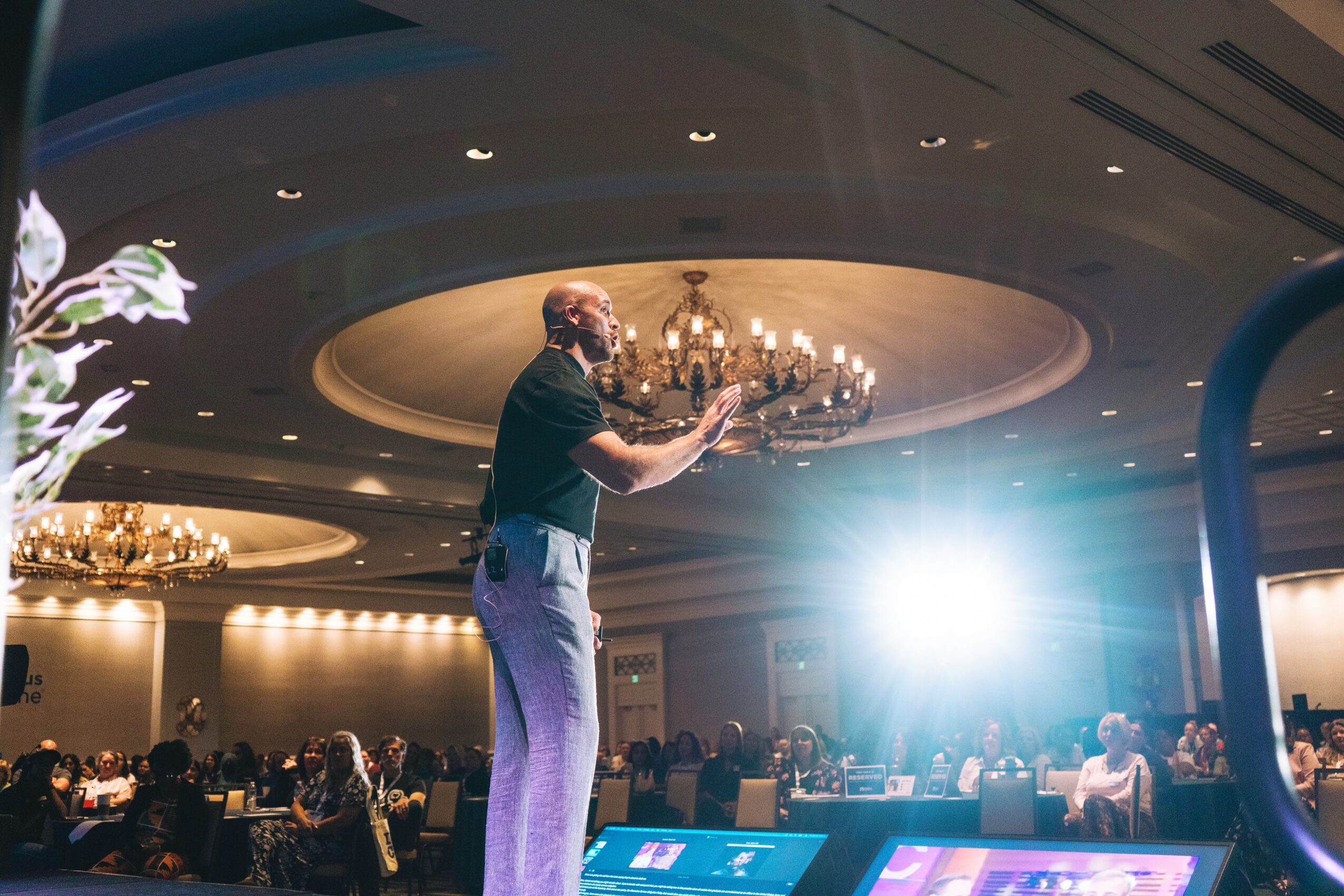 A person stands on stage in a large, elegant room with chandeliers, addressing an audience with a bright light behind them.