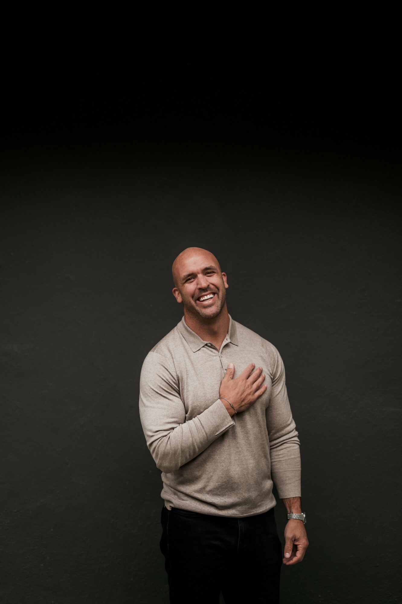A smiling man with a beard wearing a beige sweater, standing against a dark background.