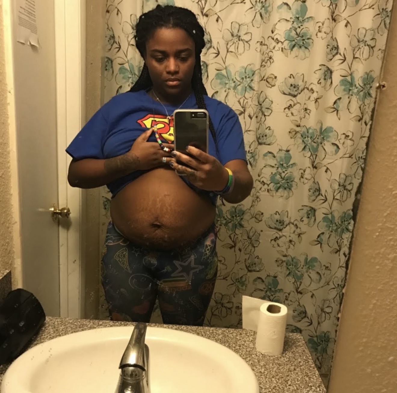 Person taking a mirror selfie in a bathroom, wearing a blue shirt with a Superman logo, holding a phone, with a large belly visible.