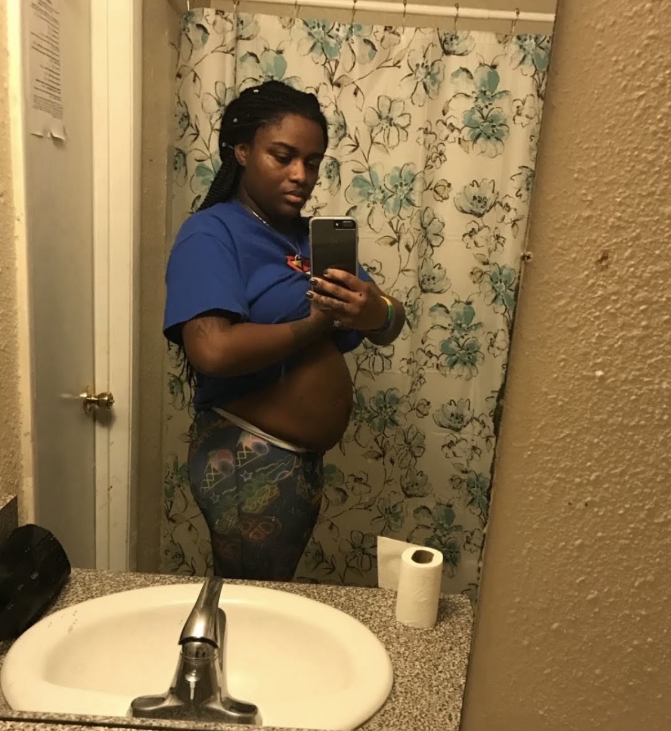 Person taking a mirror selfie in a bathroom, wearing a blue shirt and patterned pants, with a roll of toilet paper nearby.