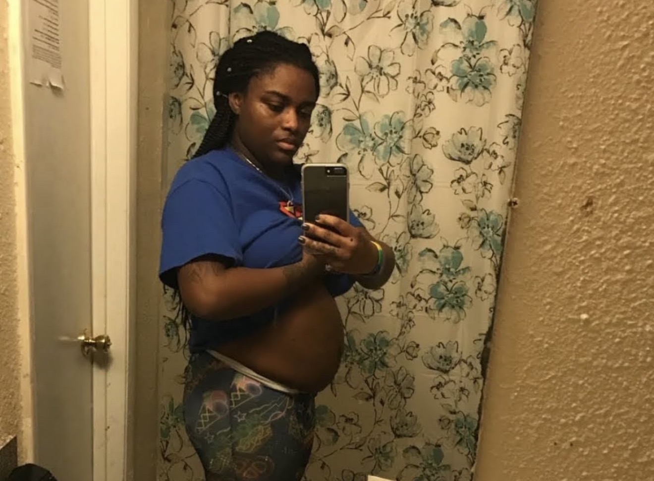Person taking a mirror selfie in a bathroom, wearing a blue shirt and patterned pants, with a floral shower curtain in the background.