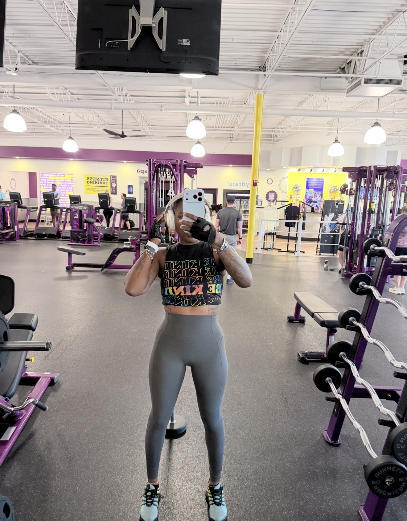 Person taking a mirror selfie in a gym, wearing a colorful crop top and gray leggings, surrounded by workout equipment.