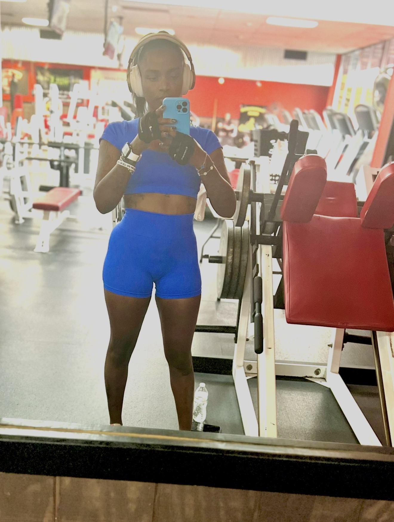 Person taking a mirror selfie in a gym, wearing blue workout clothes, with gym equipment in background.