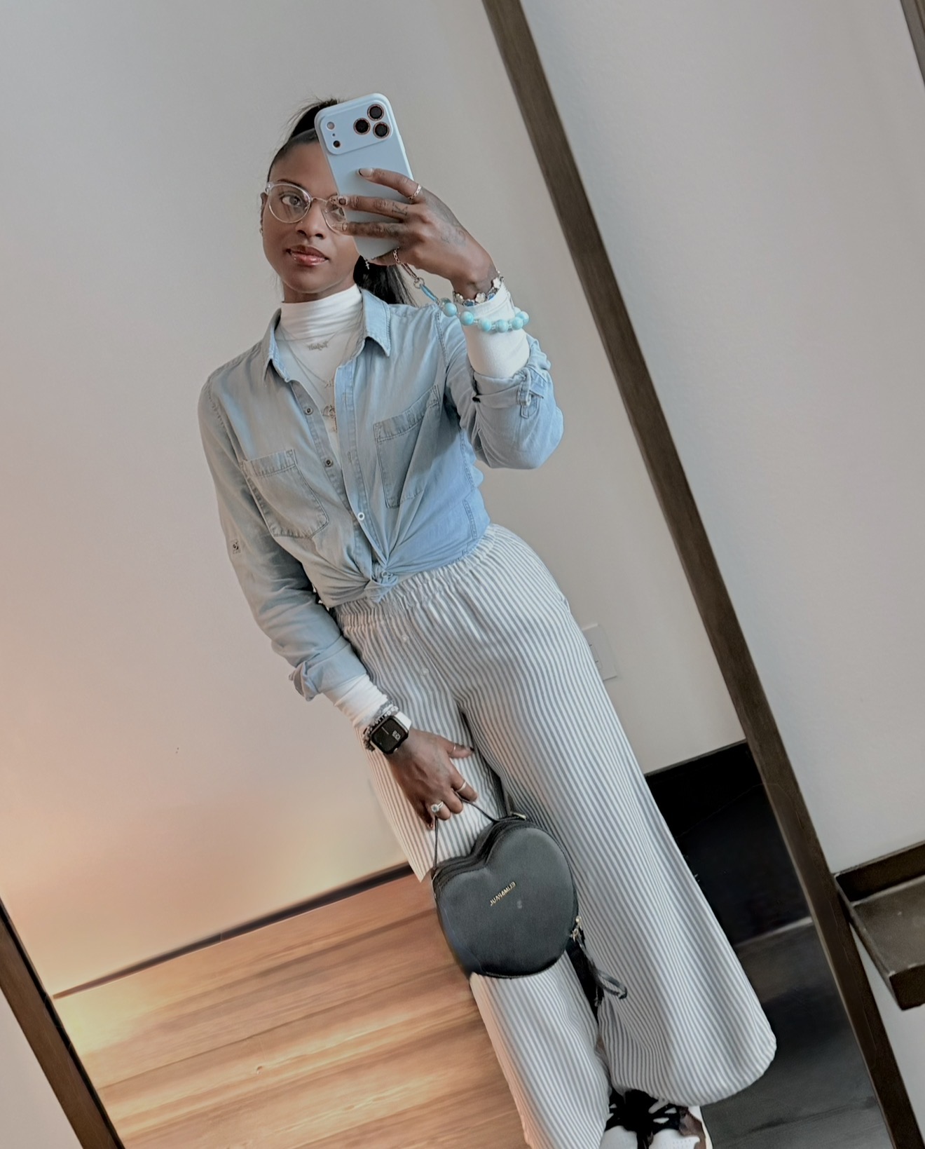 Person taking a mirror selfie with a smartphone, wearing glasses, a light blue shirt, and striped pants, holding a small bag.