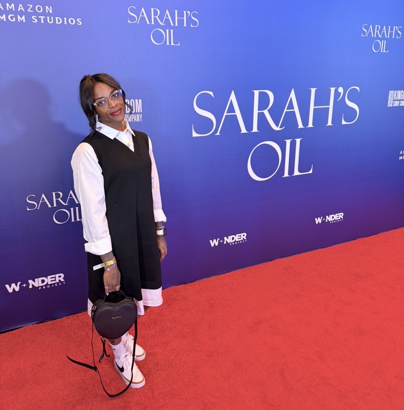 Woman standing on red carpet in front of blue backdrop with text, holding a black hat and bag, smiling.
