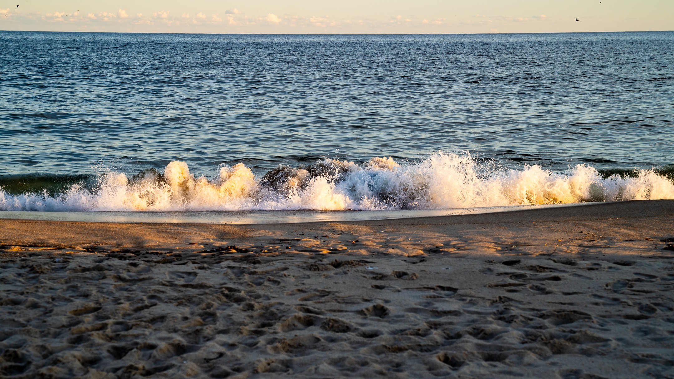 Waves crashing on a sandy beach with the ocean in the background, under a clear sky.