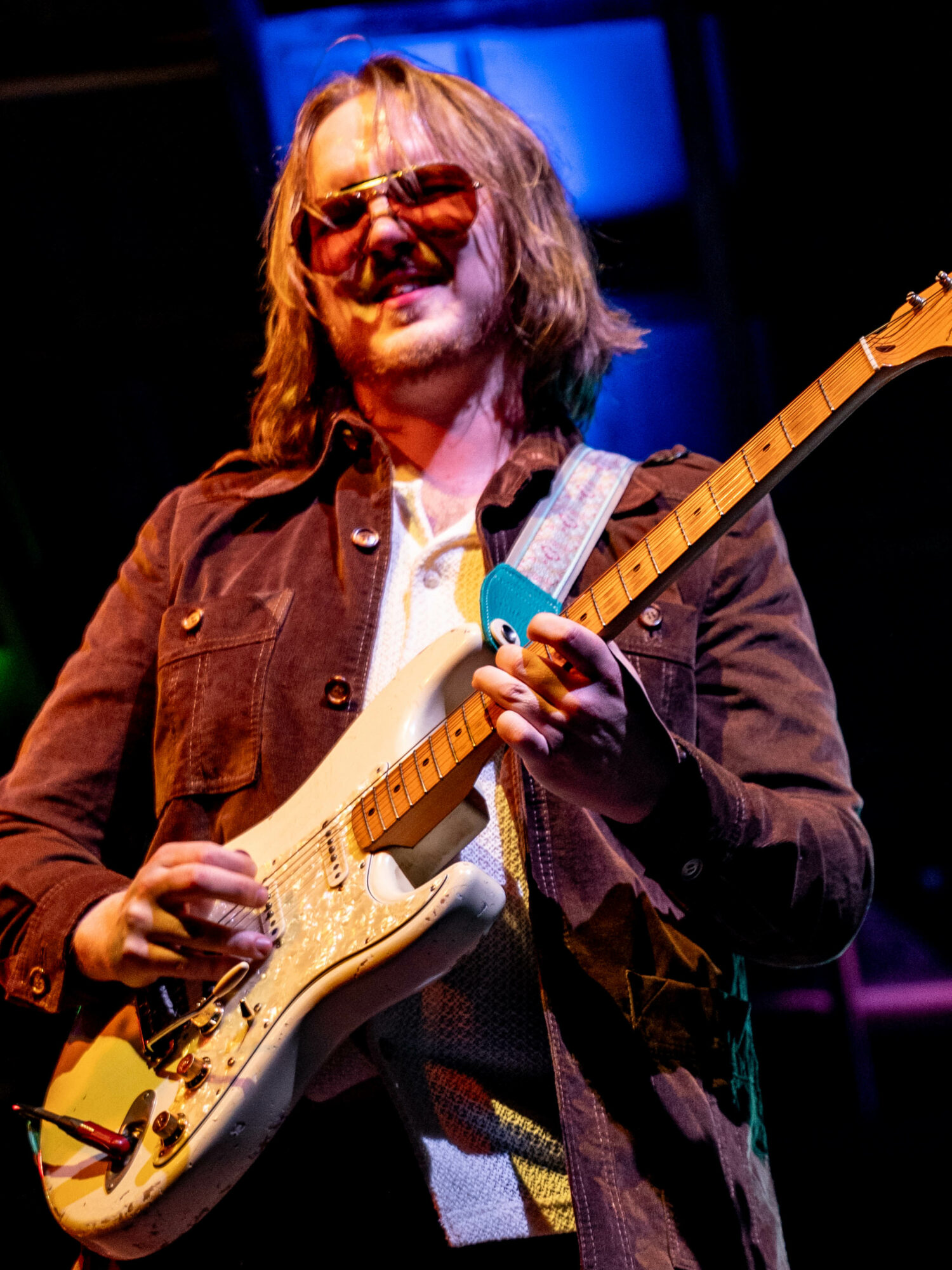 Person playing electric guitar on stage, wearing sunglasses and a brown jacket, smiling, with colorful stage lighting.