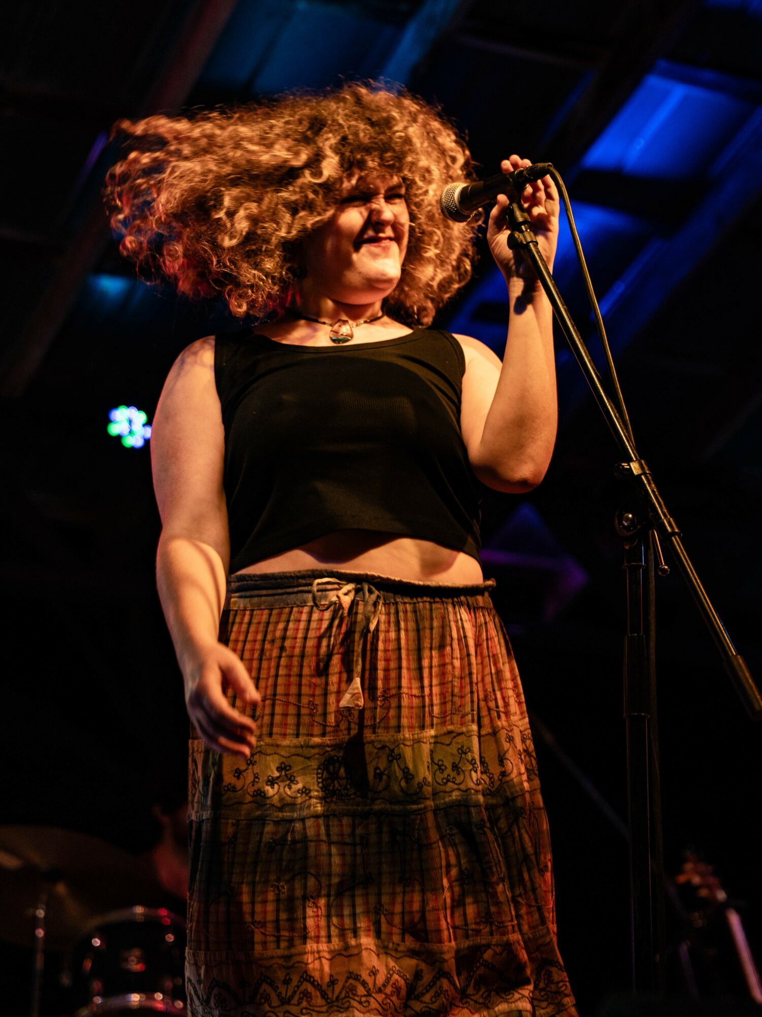 Woman with curly hair singing into a microphone on stage, smiling, wearing a black top and patterned pants.