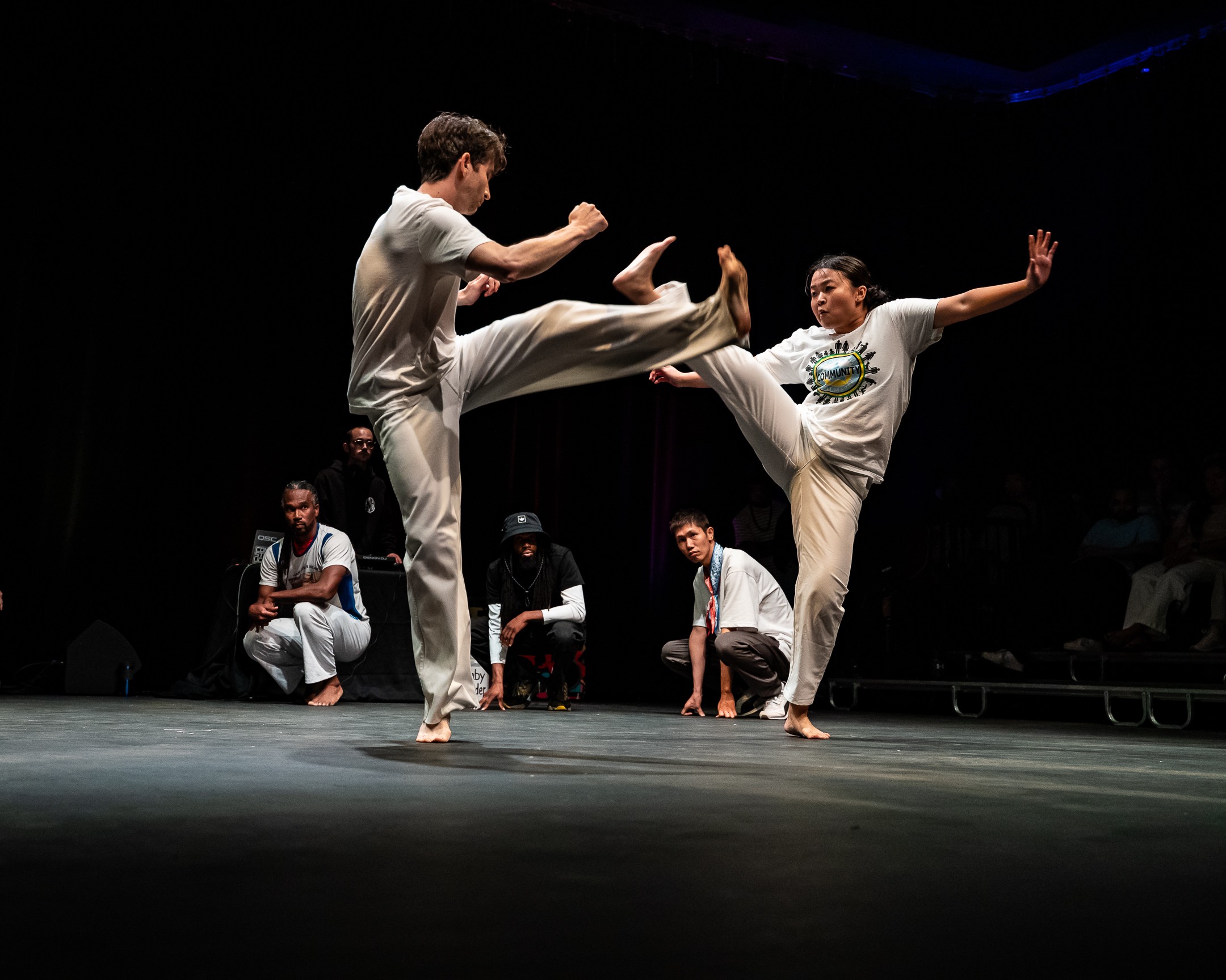 Two people perform a martial arts move on stage, with others sitting in the background, dark setting, focused lighting.