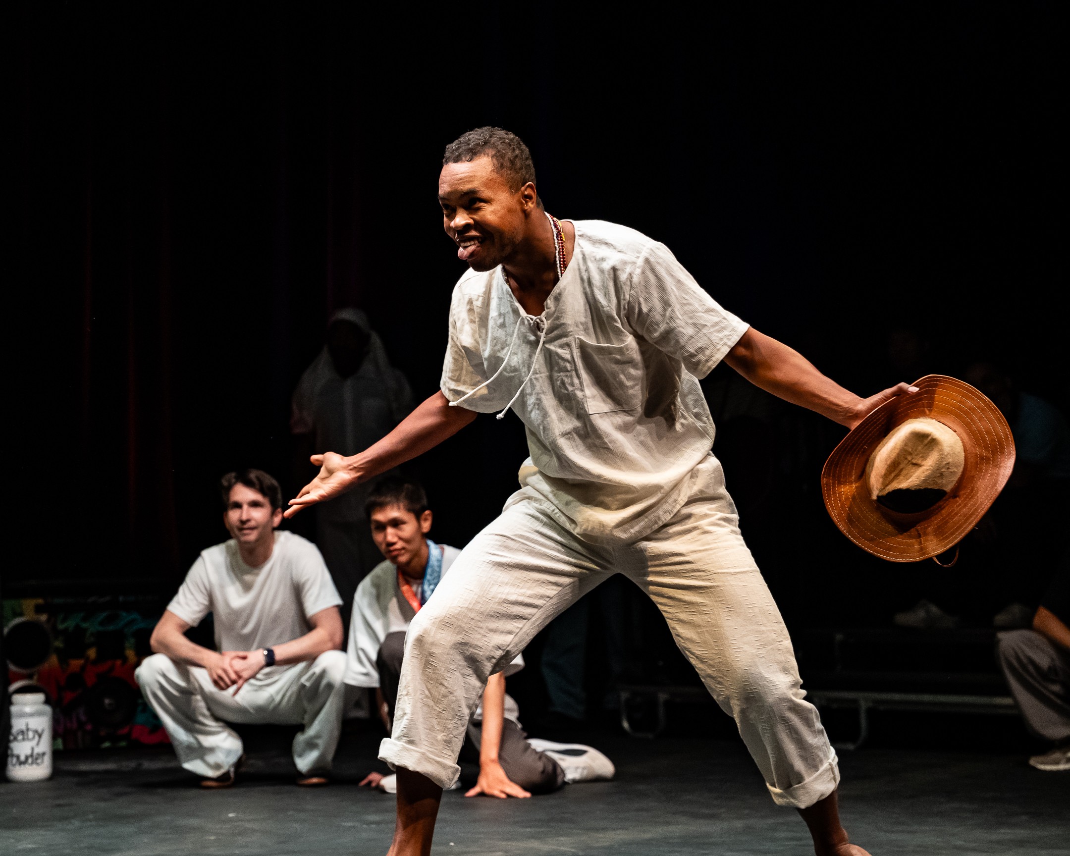 Person dancing on stage with a hat, smiling, with three seated people in the background, dark stage setting.