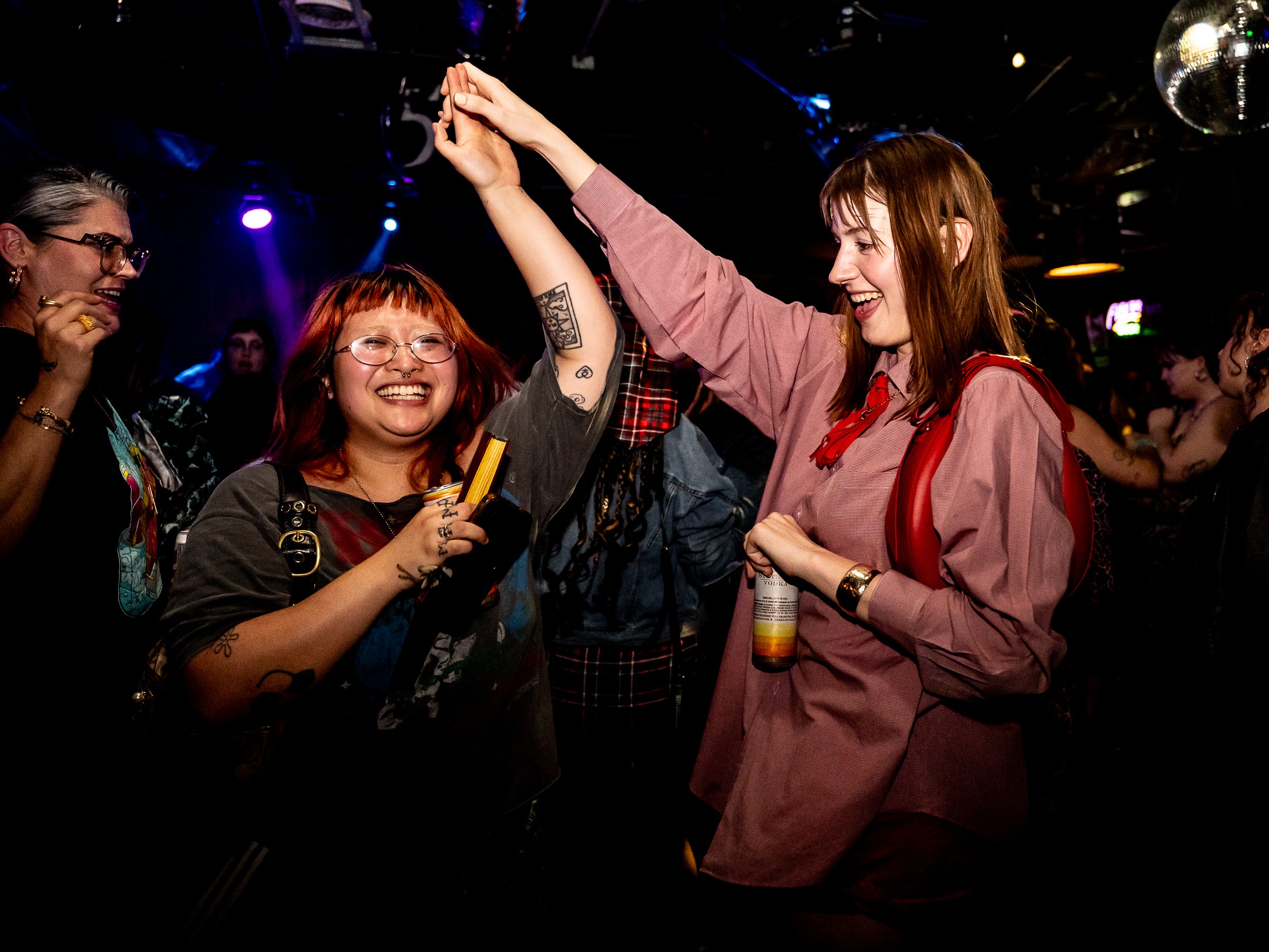 Two women dancing and smiling in a lively, dark club with colorful lights, one holding a drink and the other raising her hand.