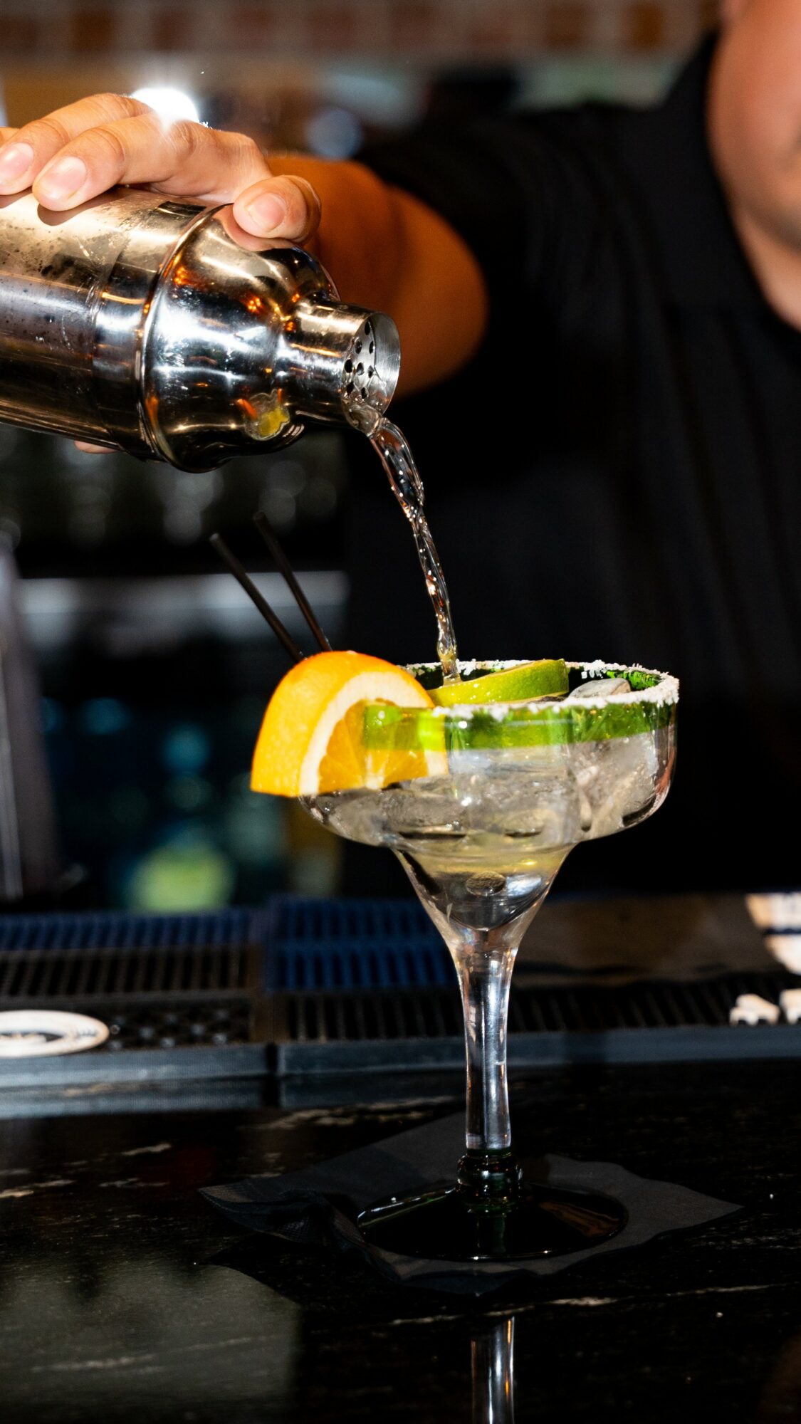 Bartender pours liquid into a cocktail glass with lemon and lime slices, against a blurred bar background.