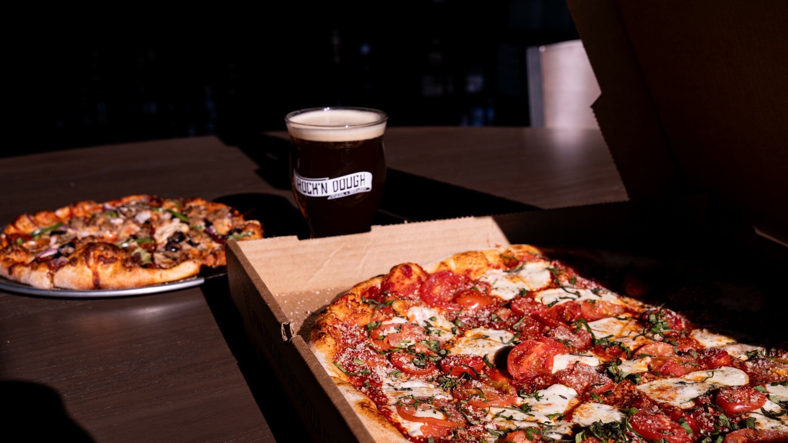 Two pizzas on a table, one in a box and one on a plate, with a glass of dark beer in the background.