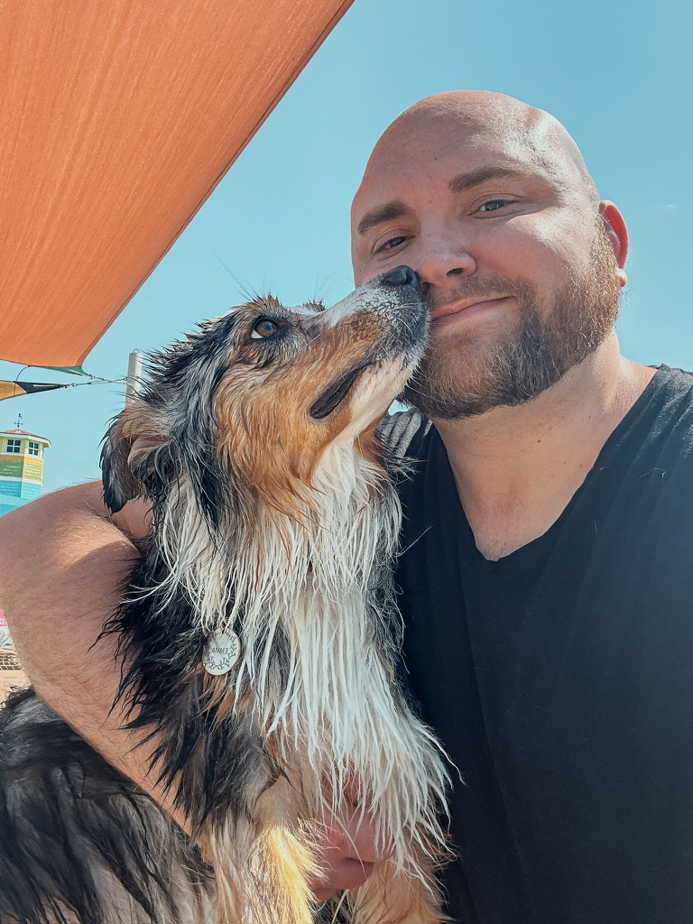 Man with a beard smiling with a wet dog outdoors against a blue sky.