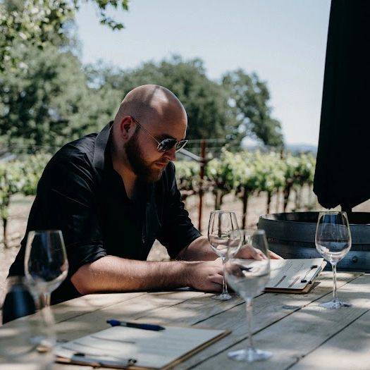 Man with sunglasses and a beard sitting at an outdoor table with wine glasses, trees, and a vineyard in the background.
