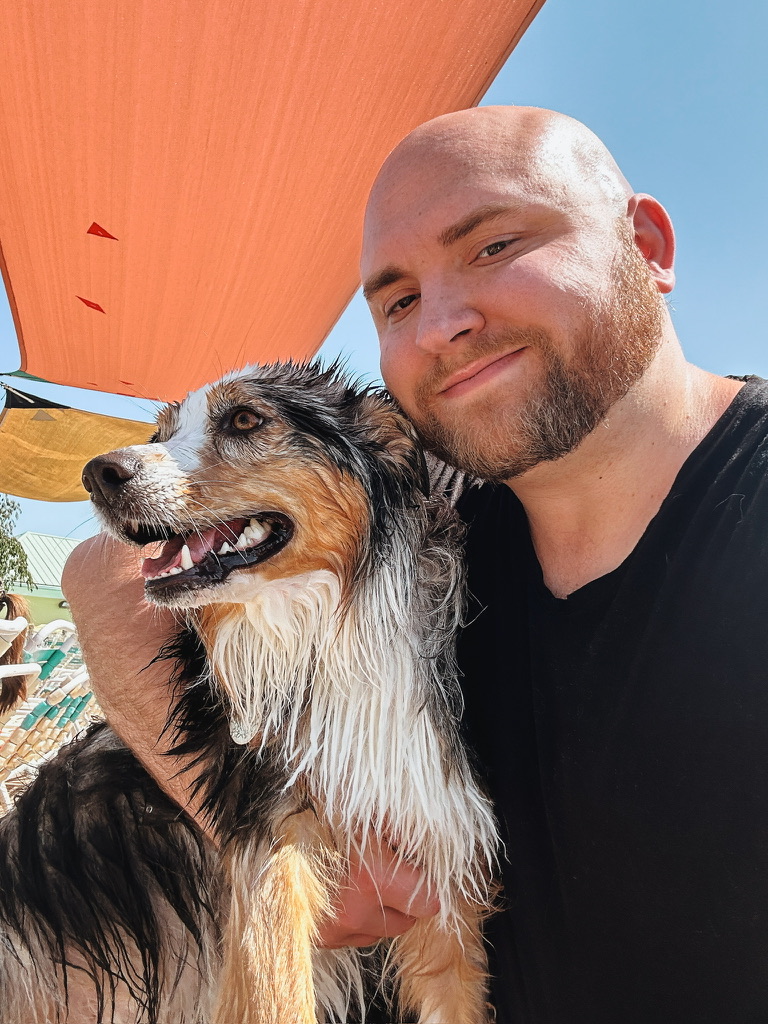 Man smiling with wet dog outdoors under a large orange shade, blue sky in background.
