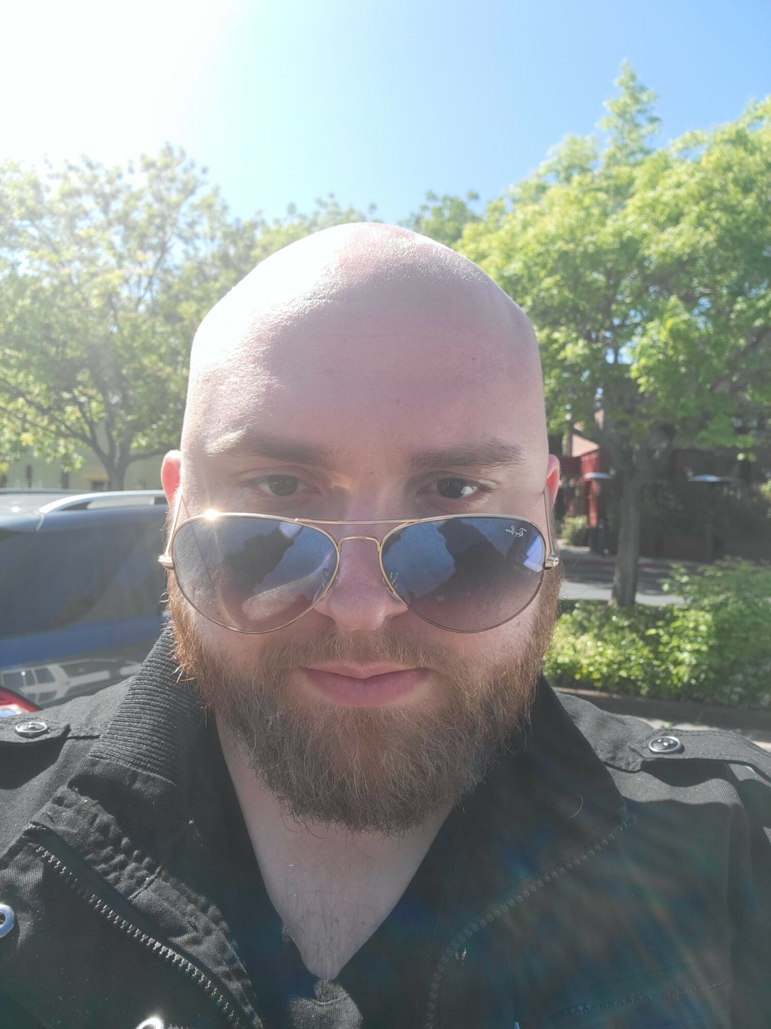 Bald man with sunglasses and beard outdoors, trees and cars in background.