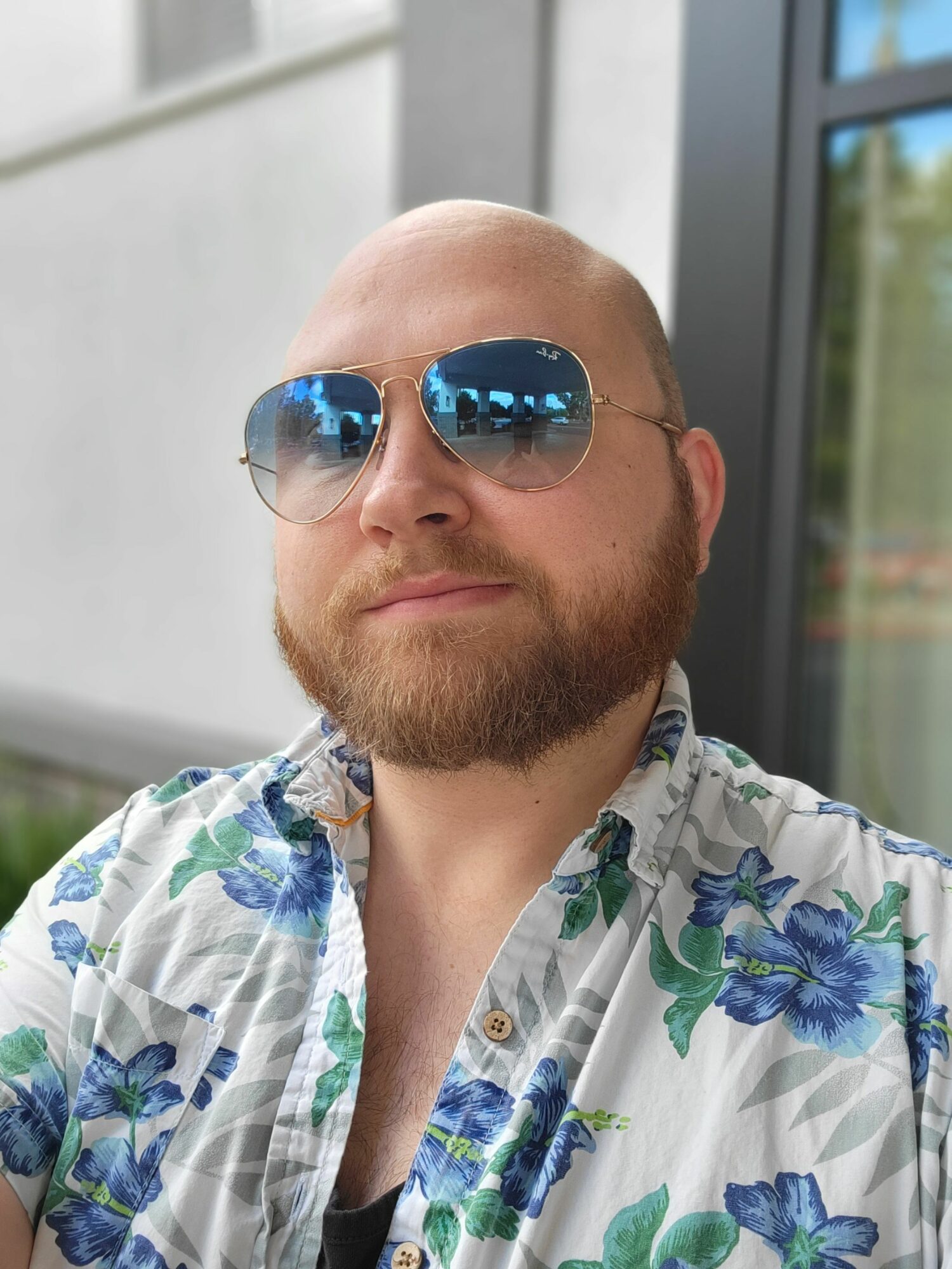 Man with a beard wearing sunglasses and a floral shirt, smiling outdoors near a building.