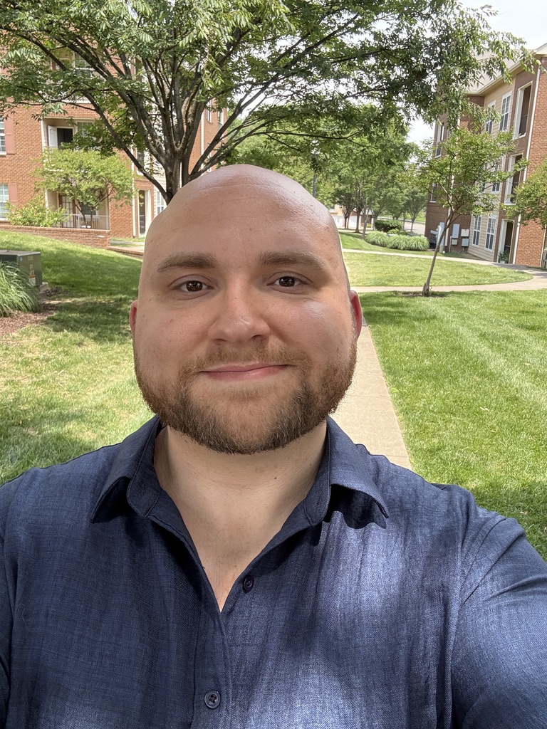 Man with a beard and short hair smiling outdoors in a park with trees and buildings in the background.