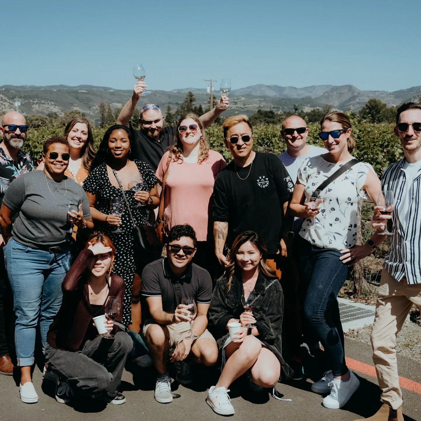 Group of people outdoors holding drinks, smiling, with mountains and blue sky in background.
