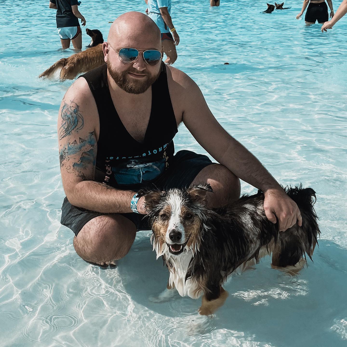 Man with sunglasses and a dog in shallow water at the beach, smiling, with other people swimming in the background.