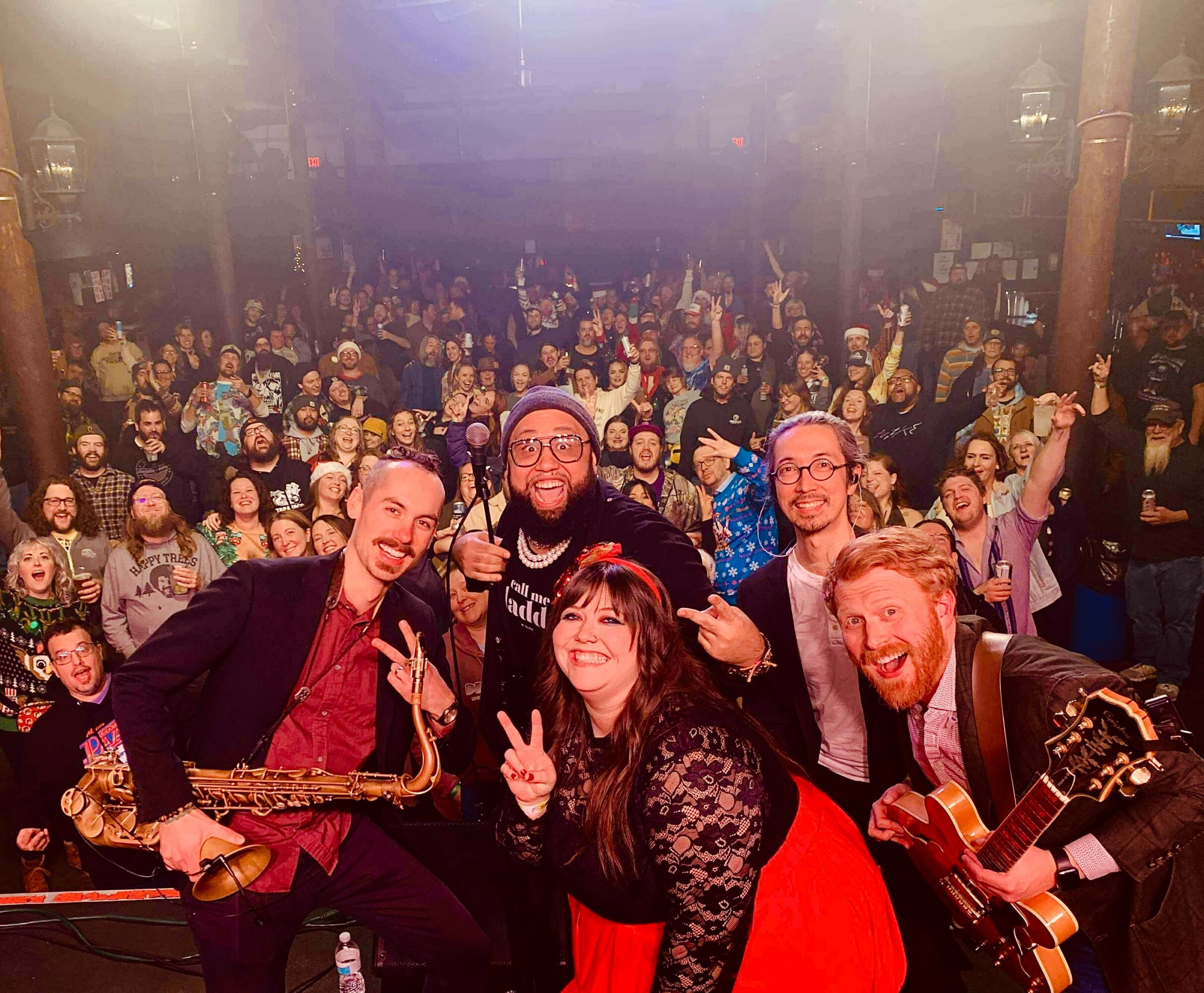 Group of five musicians on stage with a large audience behind them, some making peace signs, in a concert setting.