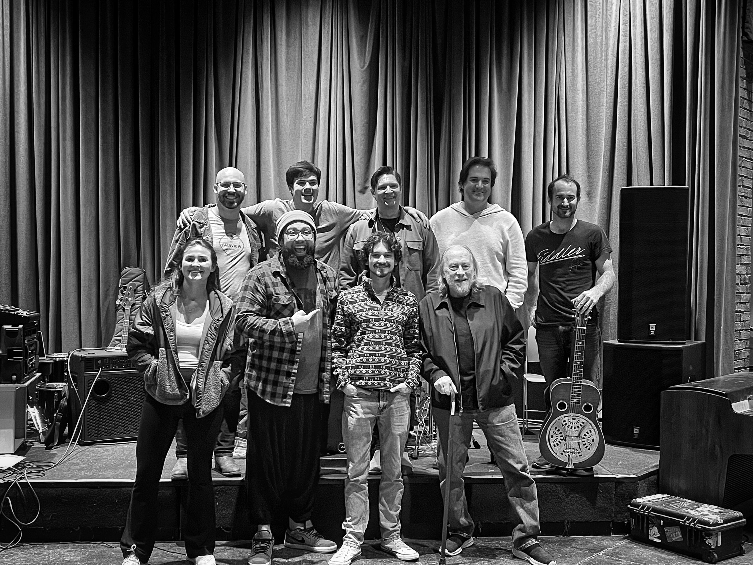 Group of ten people standing on a stage with curtains, some holding musical instruments, in black and white.