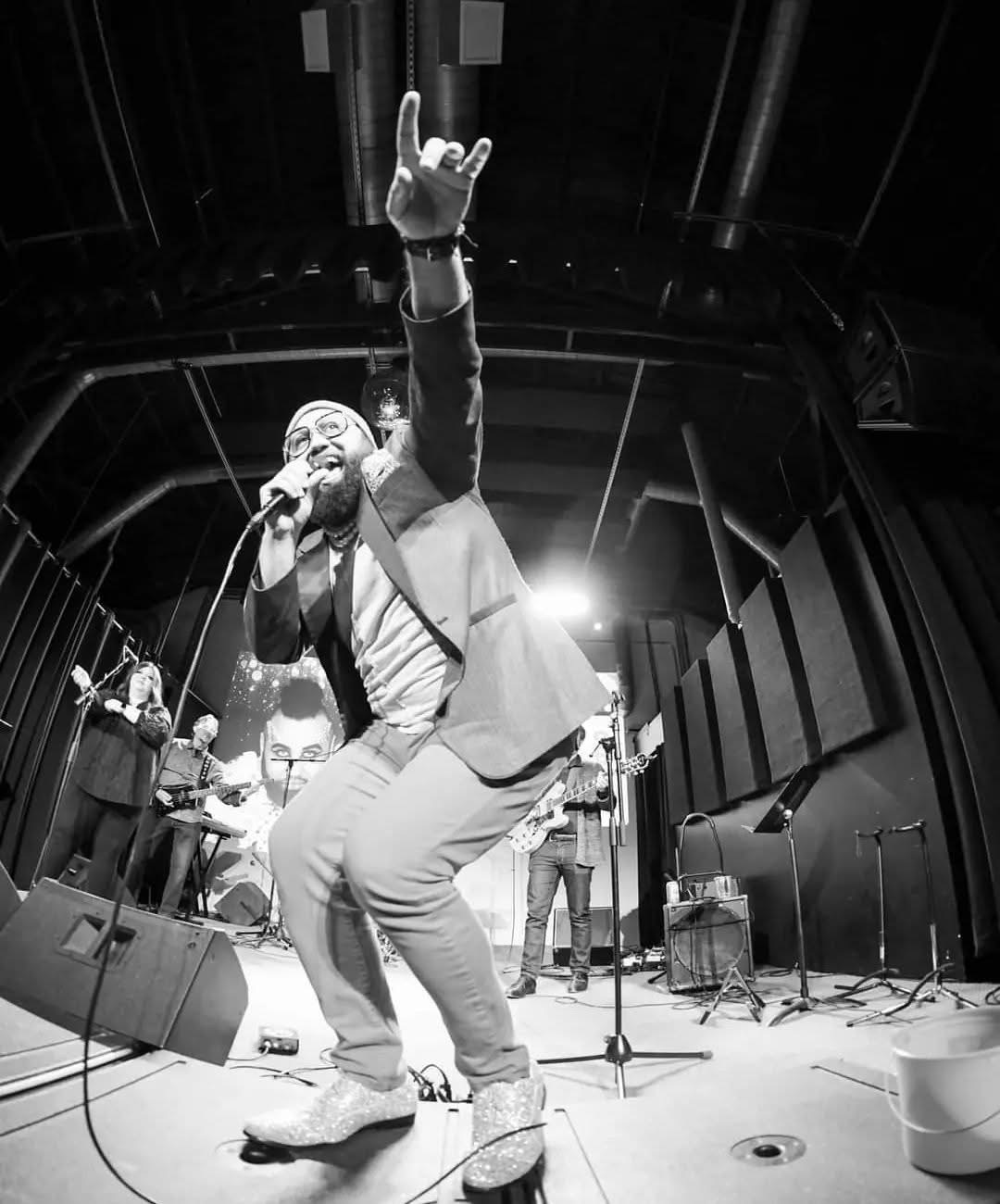 Performer singing into microphone with band on stage, raising hand, in a dark venue with stage lights.