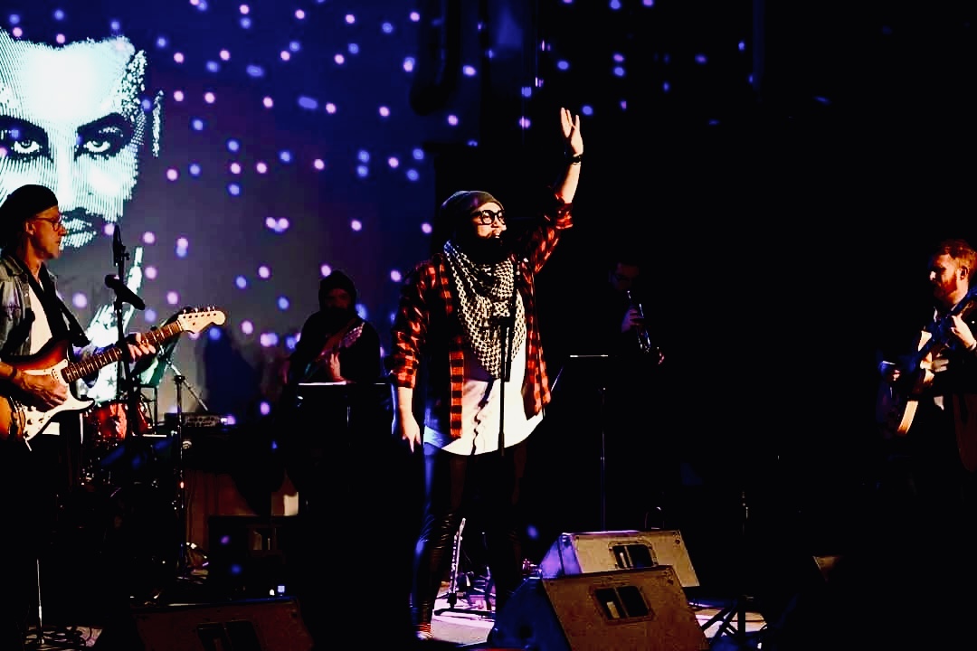 Group of musicians performing on stage with a large screen showing a face in the background, one person raising a hand.