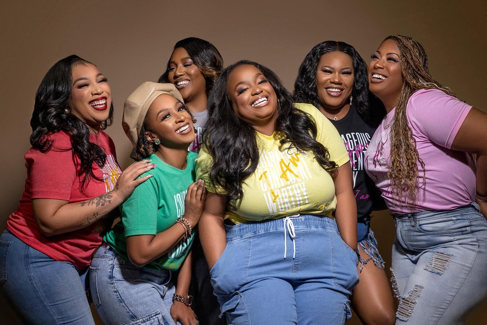 Six women smiling and posing together, wearing colorful casual clothing, against a plain background.