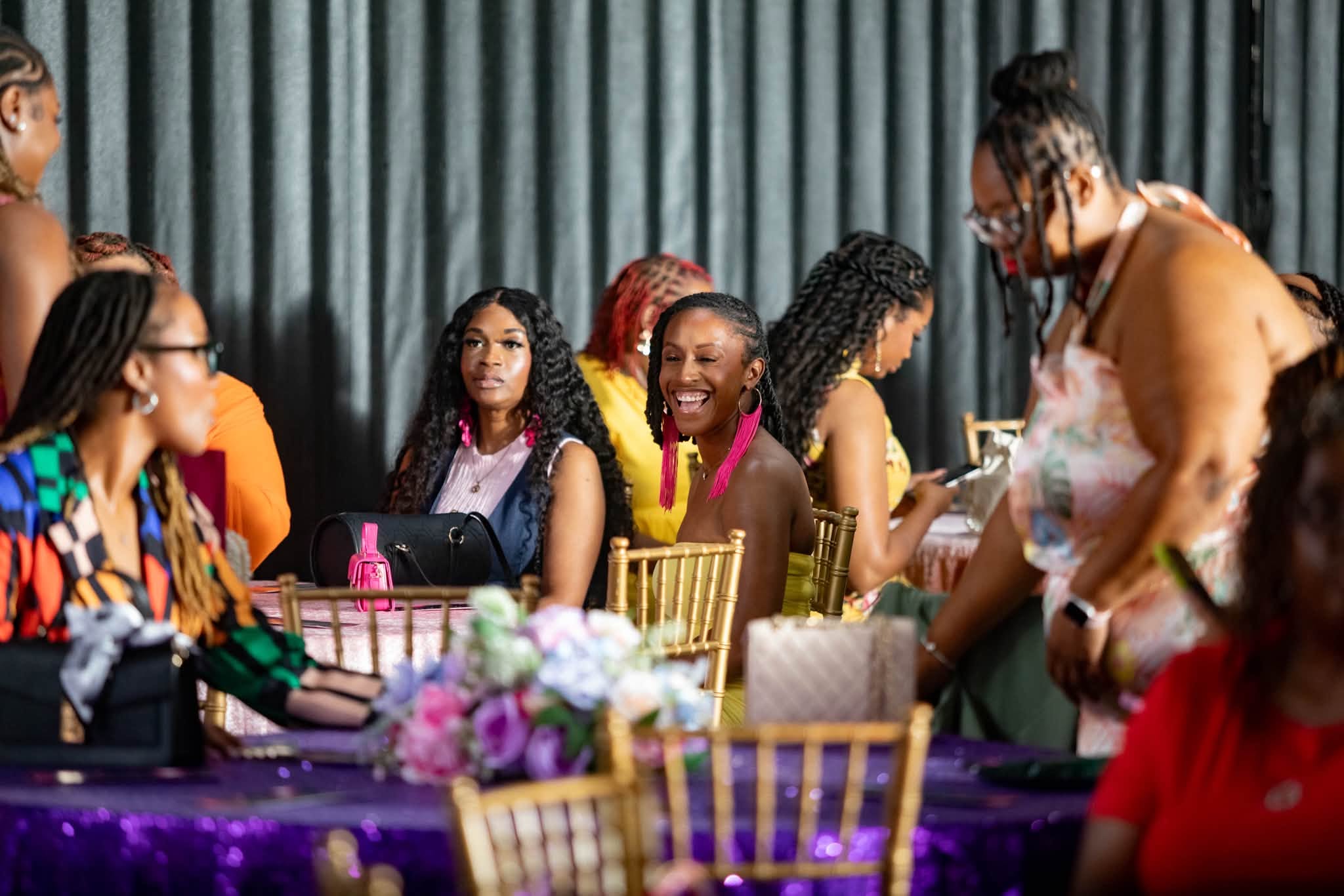 Group of women sitting at a table with purple tablecloth, smiling and talking, with a dark curtain background.