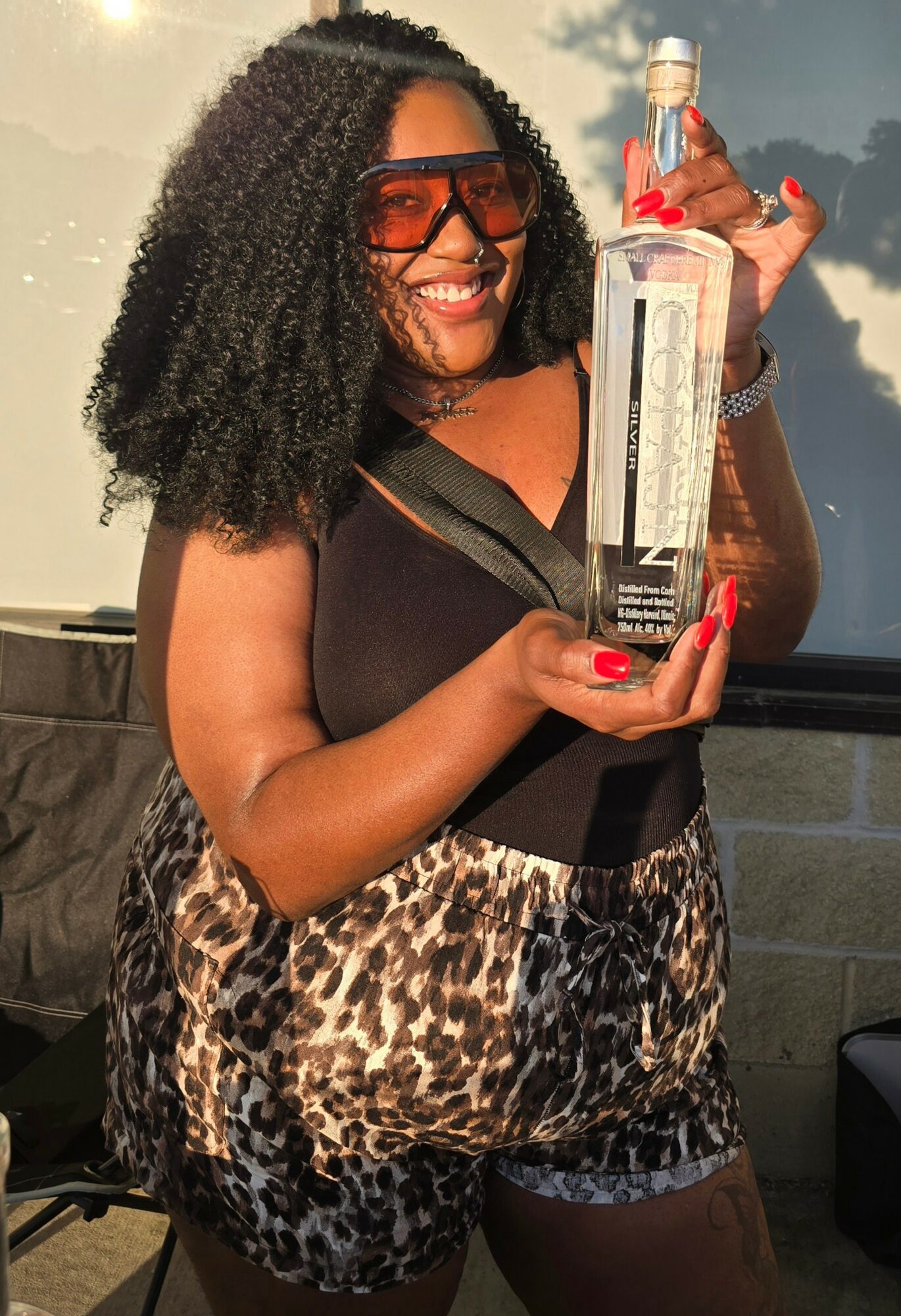 Woman with curly hair holding a bottle, smiling, wearing sunglasses and animal print shorts.
