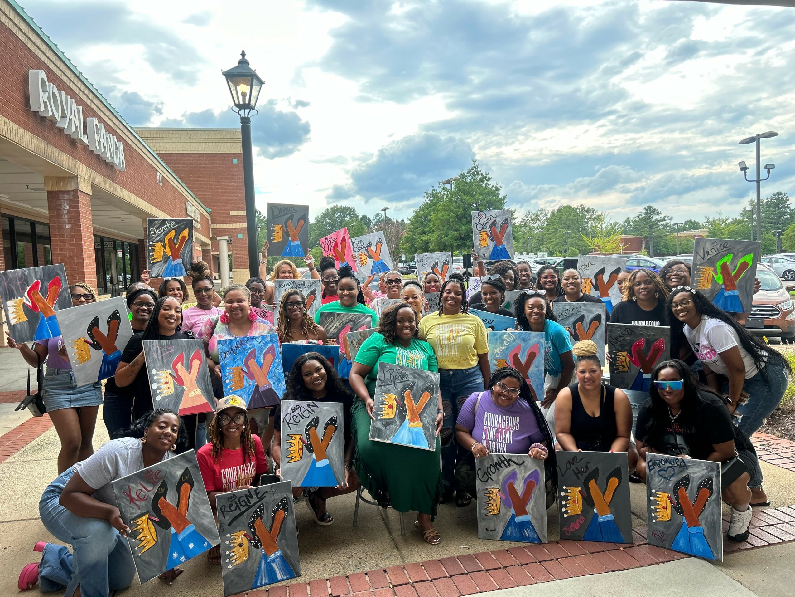 Group of people holding colorful paintings outside a building with a sign, under a partly cloudy sky.