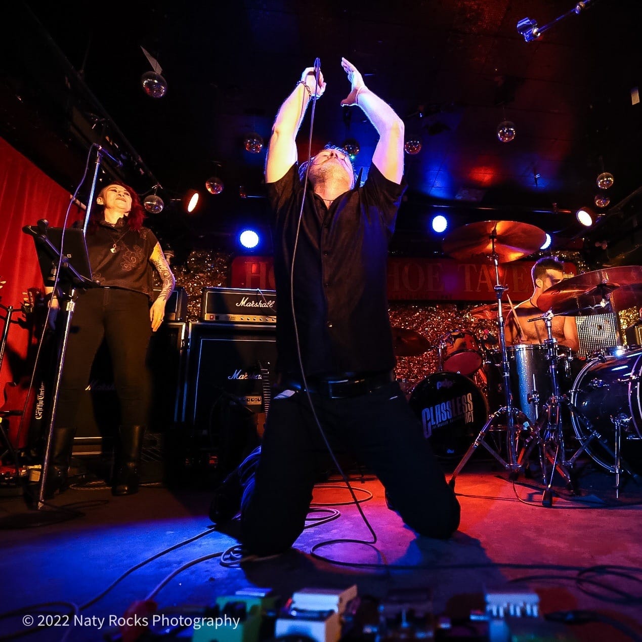 Musicians perform on stage with a singer kneeling and raising a microphone, a woman playing keyboard, and drums in background.