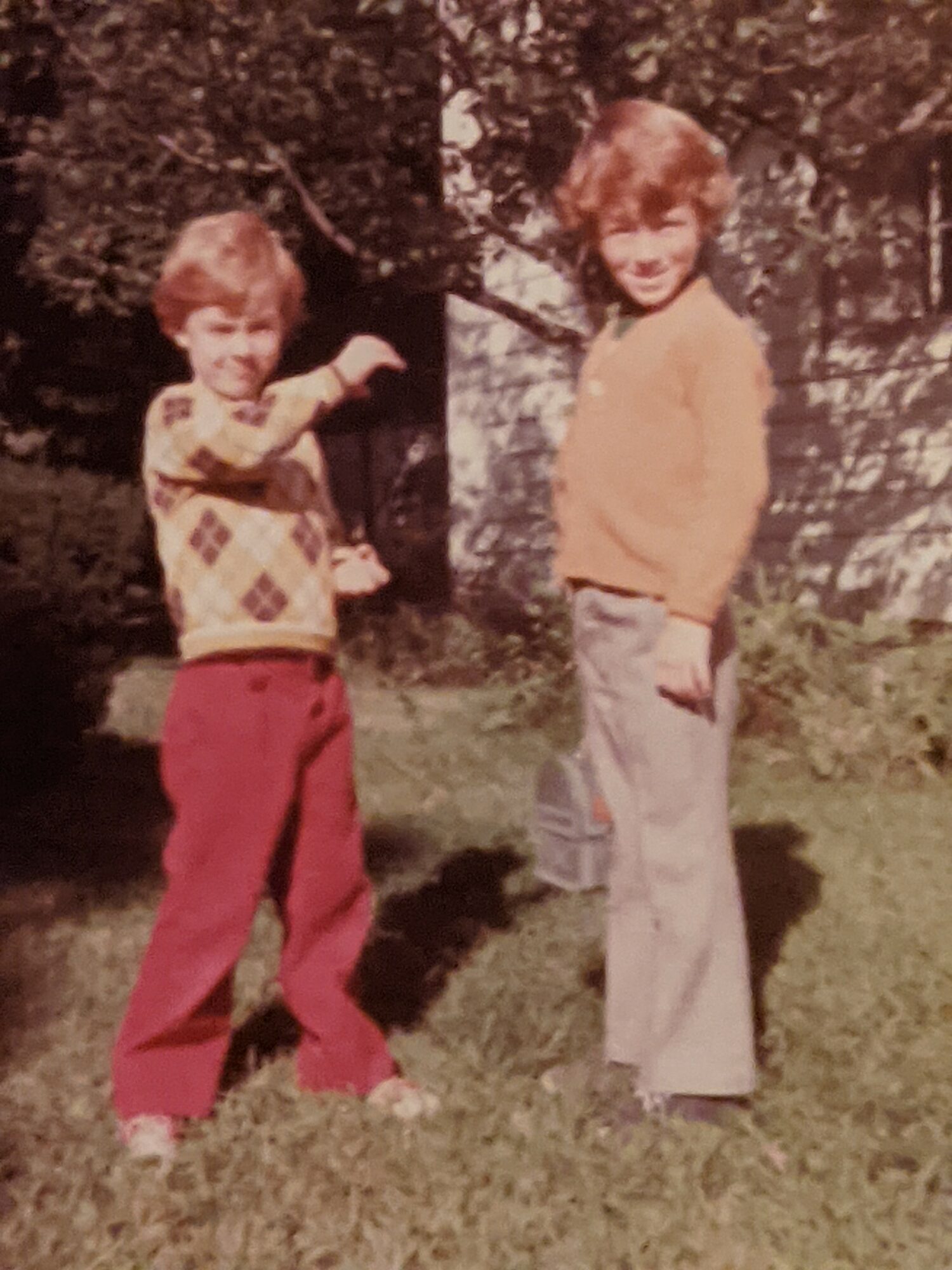 Two children standing outdoors on grass, with trees and a building in the background, smiling at the camera.
