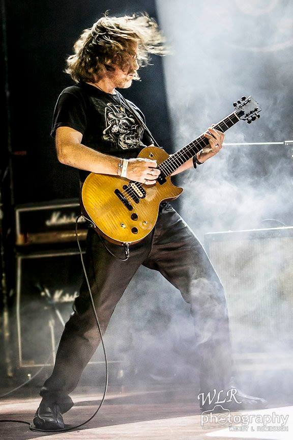 Person playing an electric guitar on stage with smoke and stage lights, hair flying, wearing a black t-shirt and dark pants.
