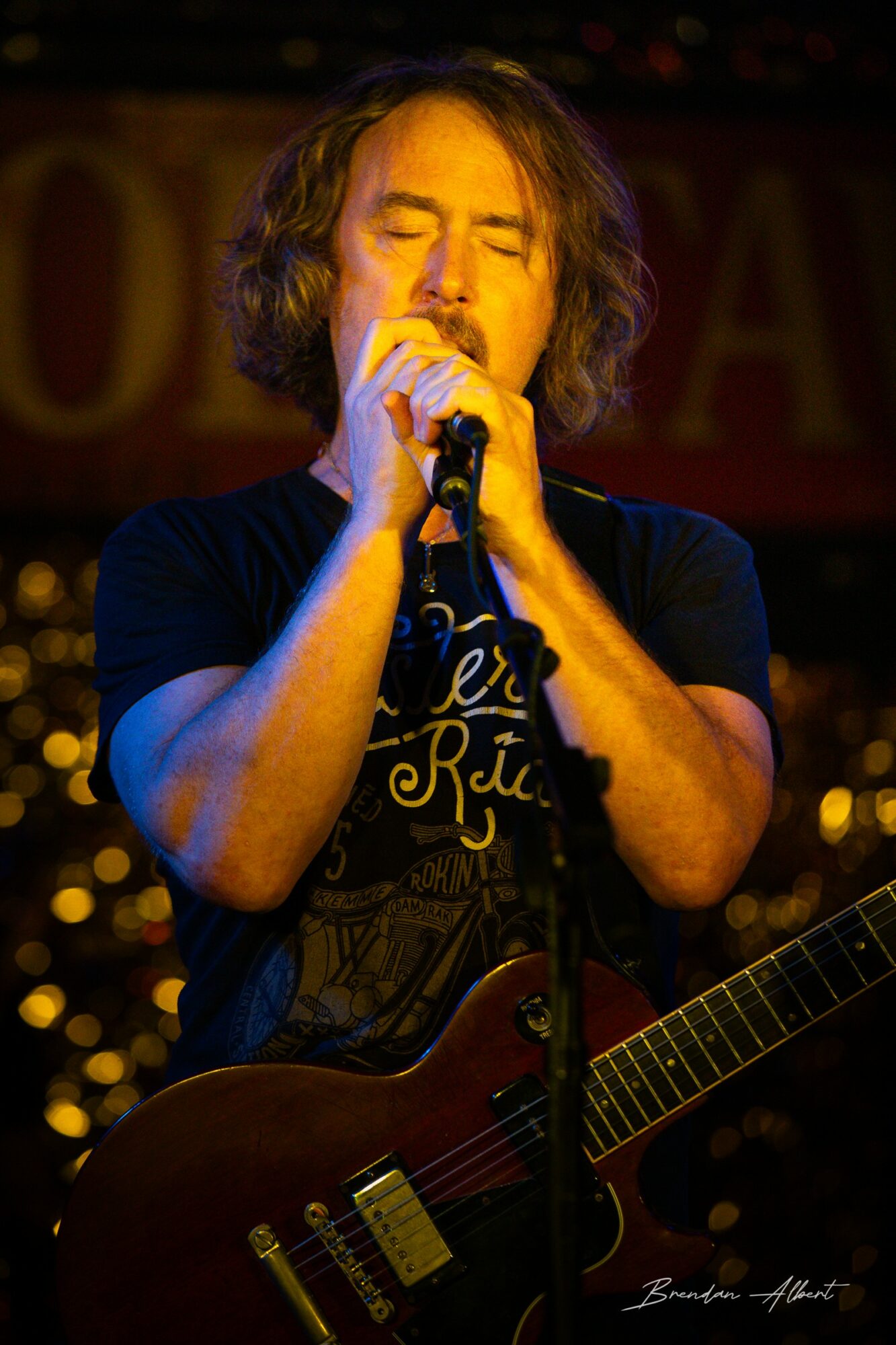 Person with shoulder-length hair singing into a microphone, holding a guitar, with closed eyes and a focused expression.