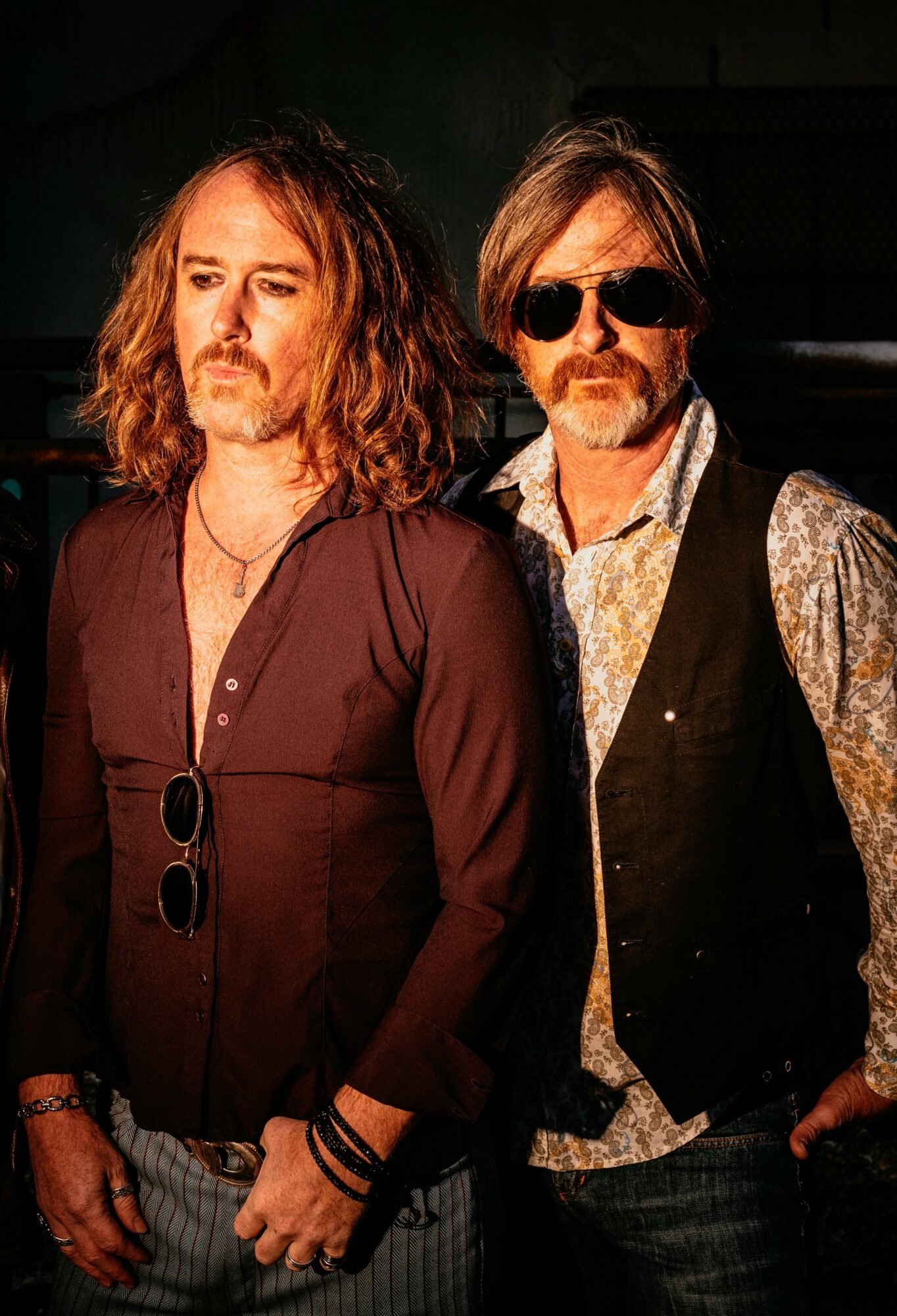 Two men with long hair and sunglasses stand side by side, one wearing a dark shirt and the other a patterned shirt and vest.