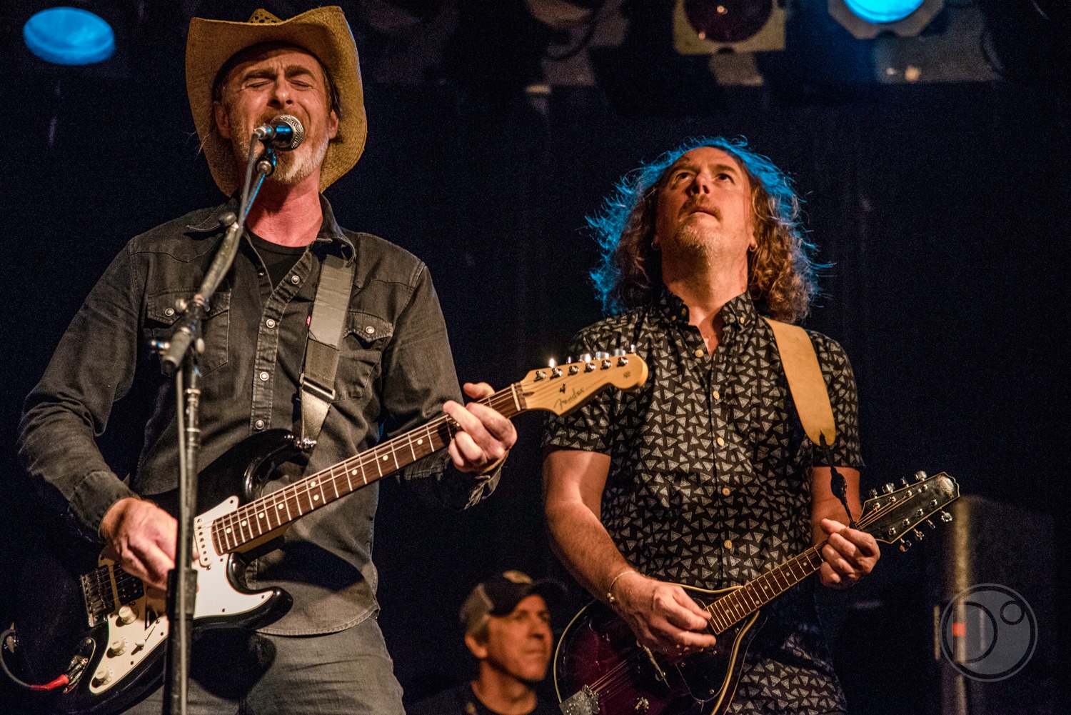 Two musicians perform on stage, one singing and playing guitar, the other playing a bass guitar, with stage lights overhead.