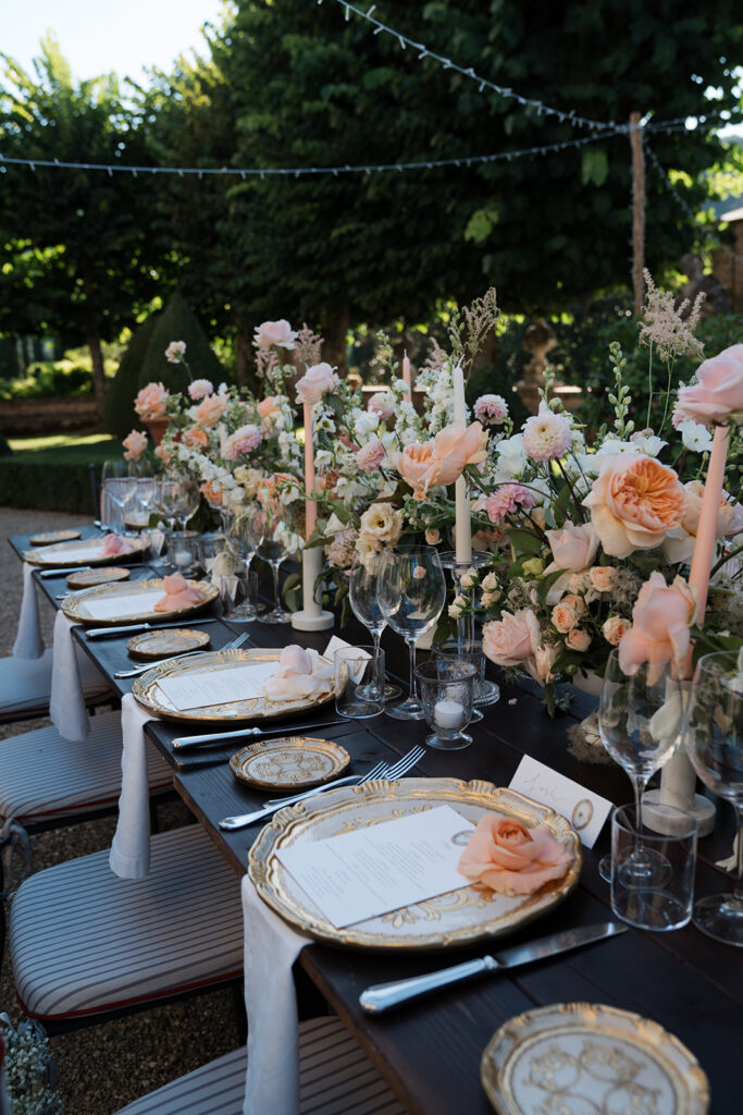 Outdoor table set with floral centerpiece, plates, glasses, and chairs, under trees with string lights, during daytime.
