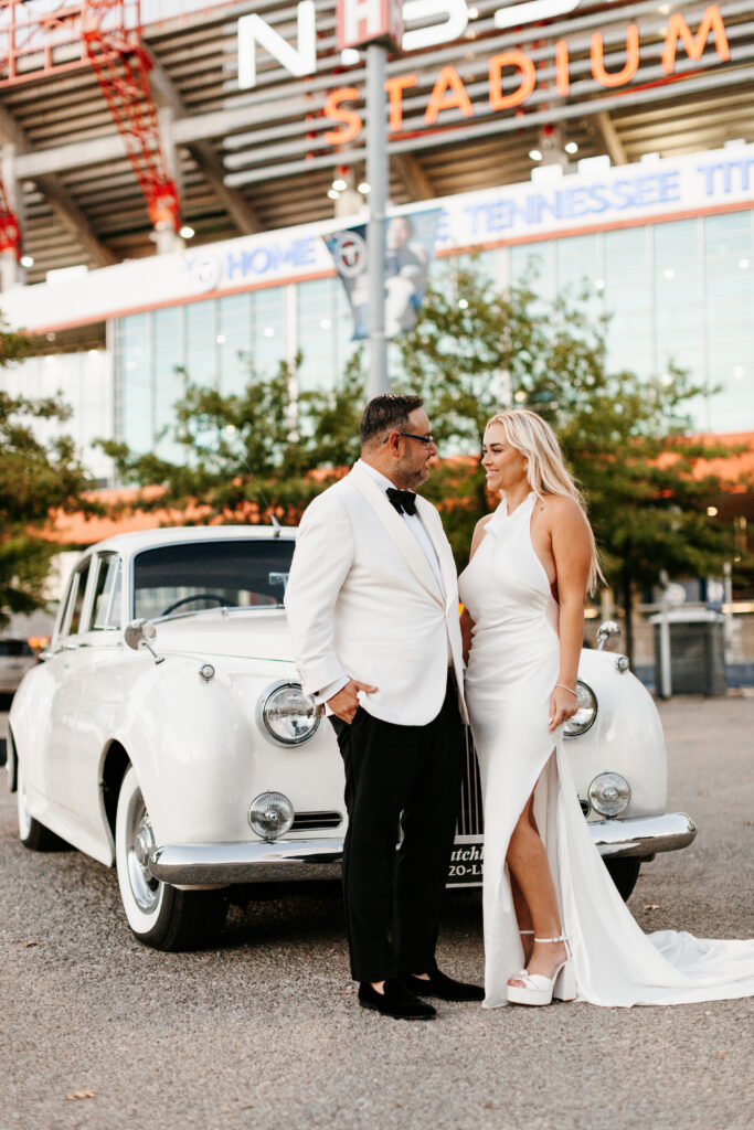 A couple in formal attire stands in front of a vintage white car outside a stadium, smiling at each other.