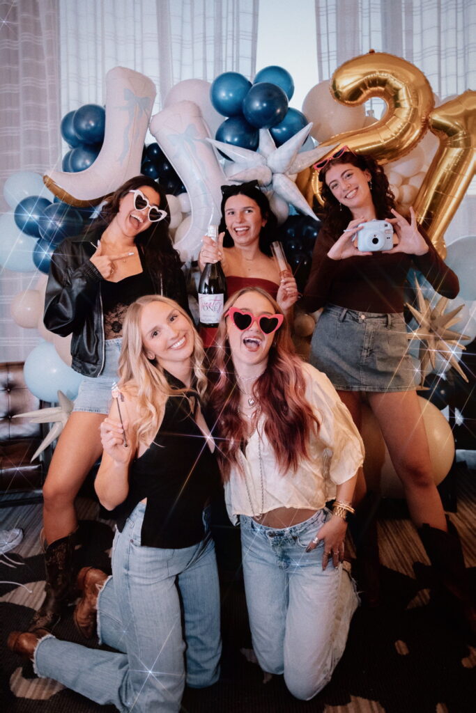 Group of five women celebrating with balloons and decorations, smiling and posing for the camera.