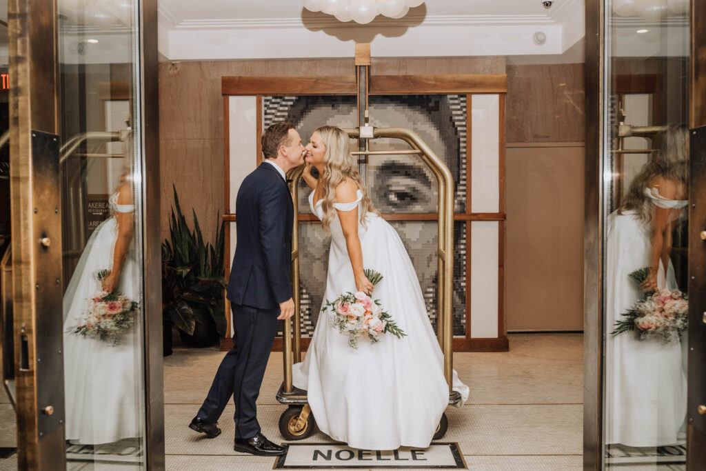 A bride and groom stand close together in a hotel lobby, with a large eye mural behind them, and reflections in glass doors.