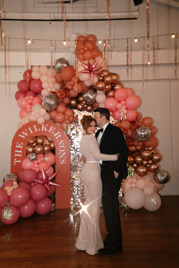 A couple in formal attire stands in front of a balloon arch with pink, gold, and silver balloons, and a pink sign reading 'The Wilkers & Sons'.