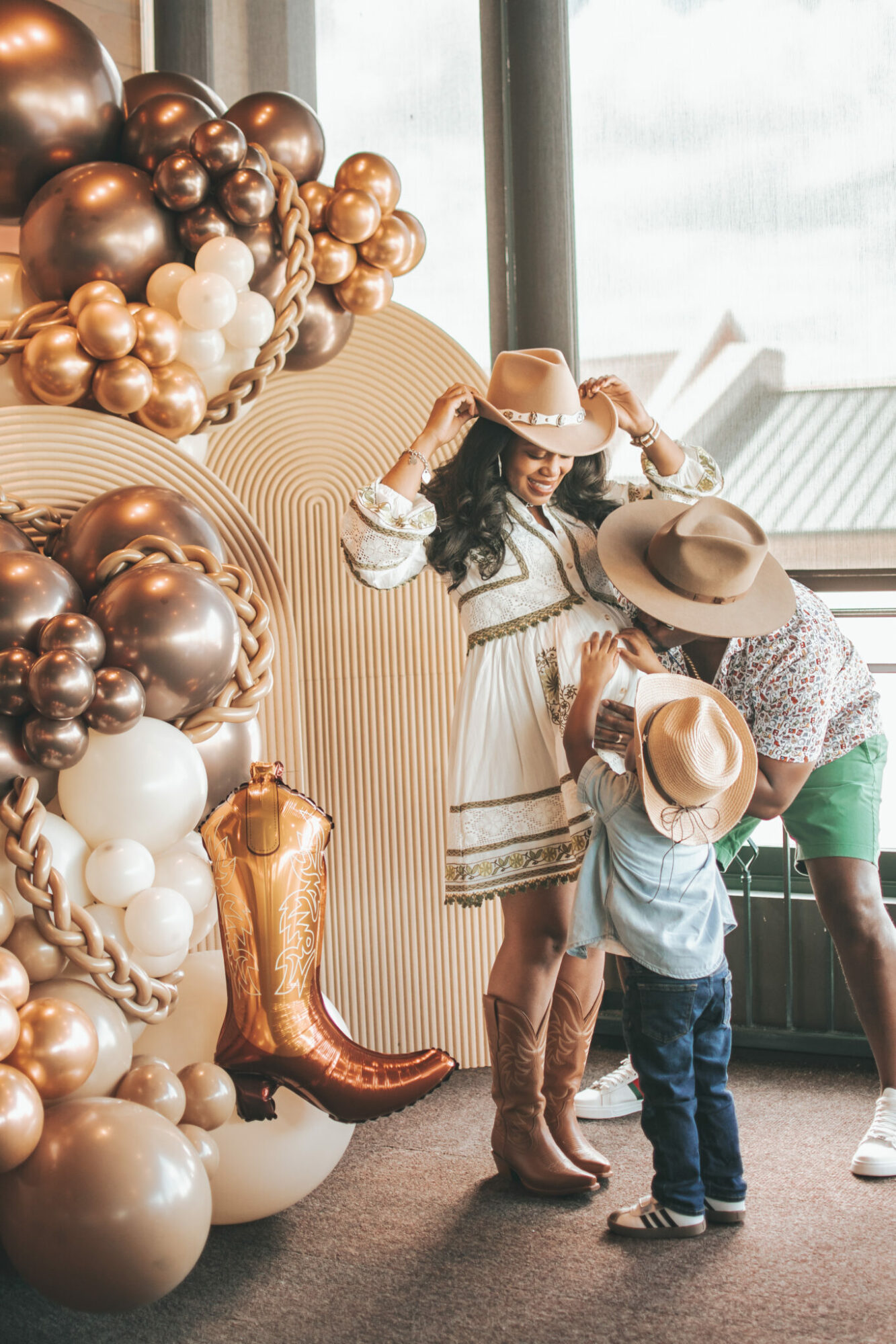 Woman in a dress and hat holding a hat, children wearing hats, balloons, and cowboy boots near a window.