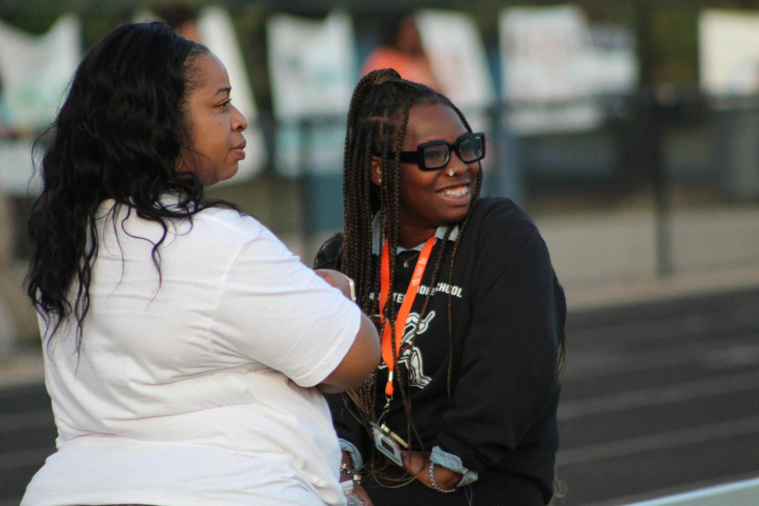 Two women sitting outdoors, one with long curly hair and the other with braided hair and sunglasses, smiling and talking.