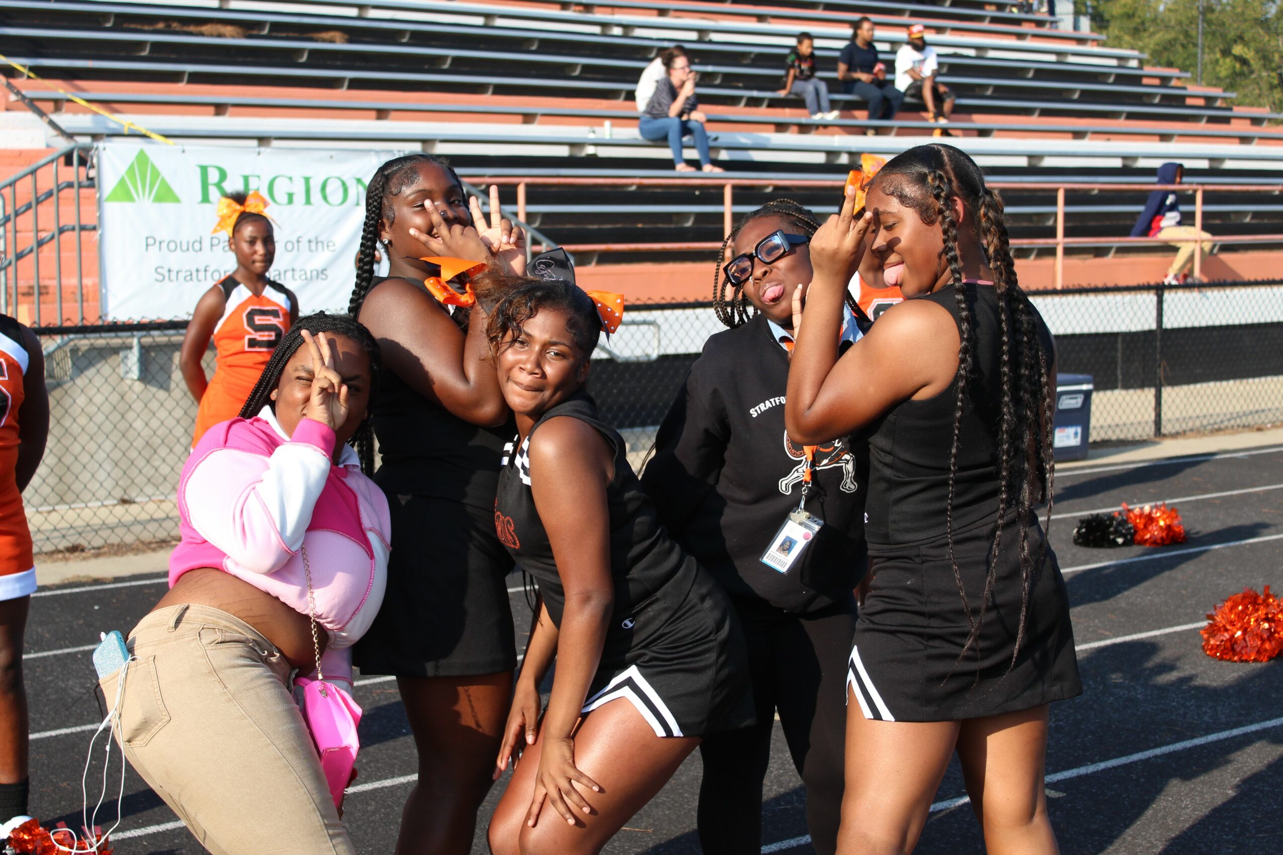 Group of young women in black athletic outfits standing on a sports field, some smiling and talking, with bleachers and spectators in background.